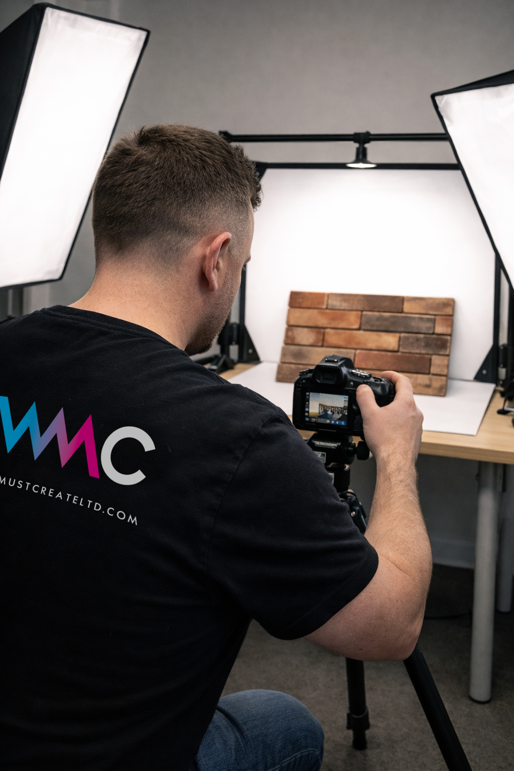 A man in a black t-shirt taking a photo of a bricks in a photography studio with two softbox lights.