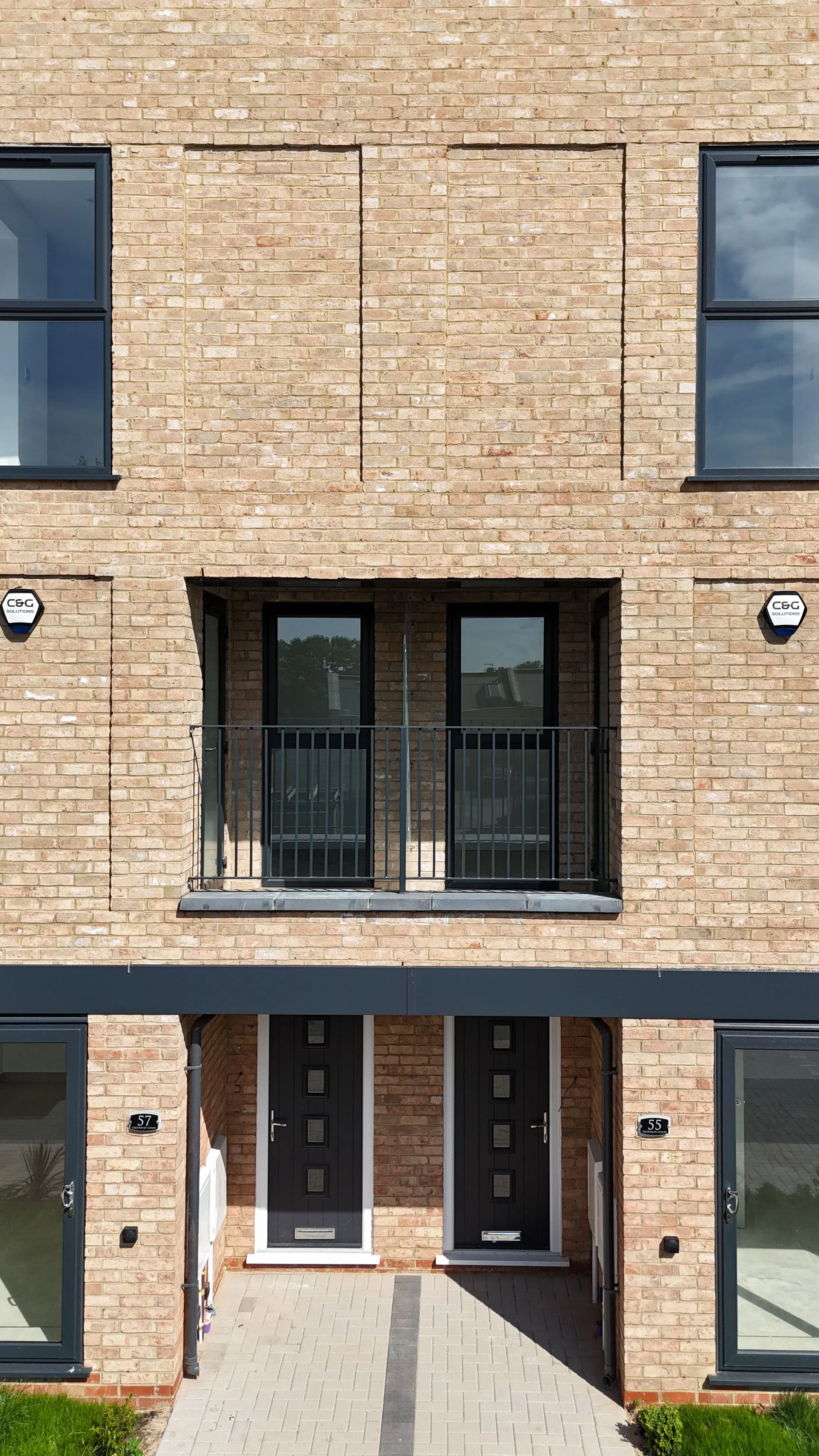 Front view of a modern brick apartment building with two black front doors, a small balcony with a black metal railing, and large windows. The building has a gray awning above the entrance and house numbers 57 and 55.