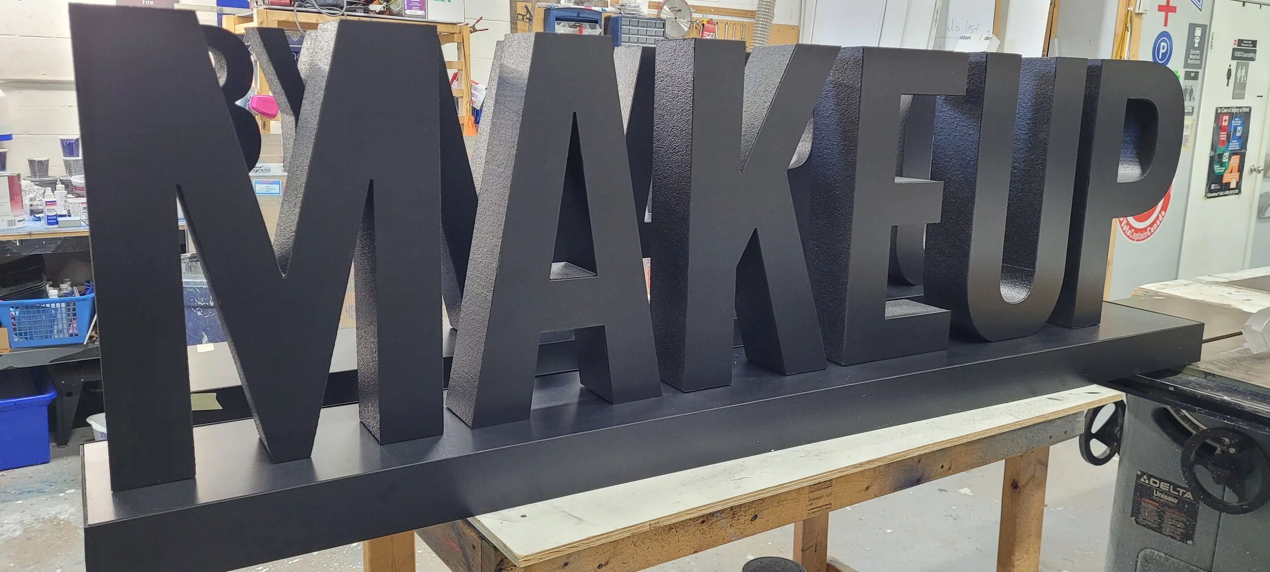 Large black 3D letters spelling 'MAKEUP' arranged on a flat black surface in a workshop or store, with tools and supplies visible in the background.