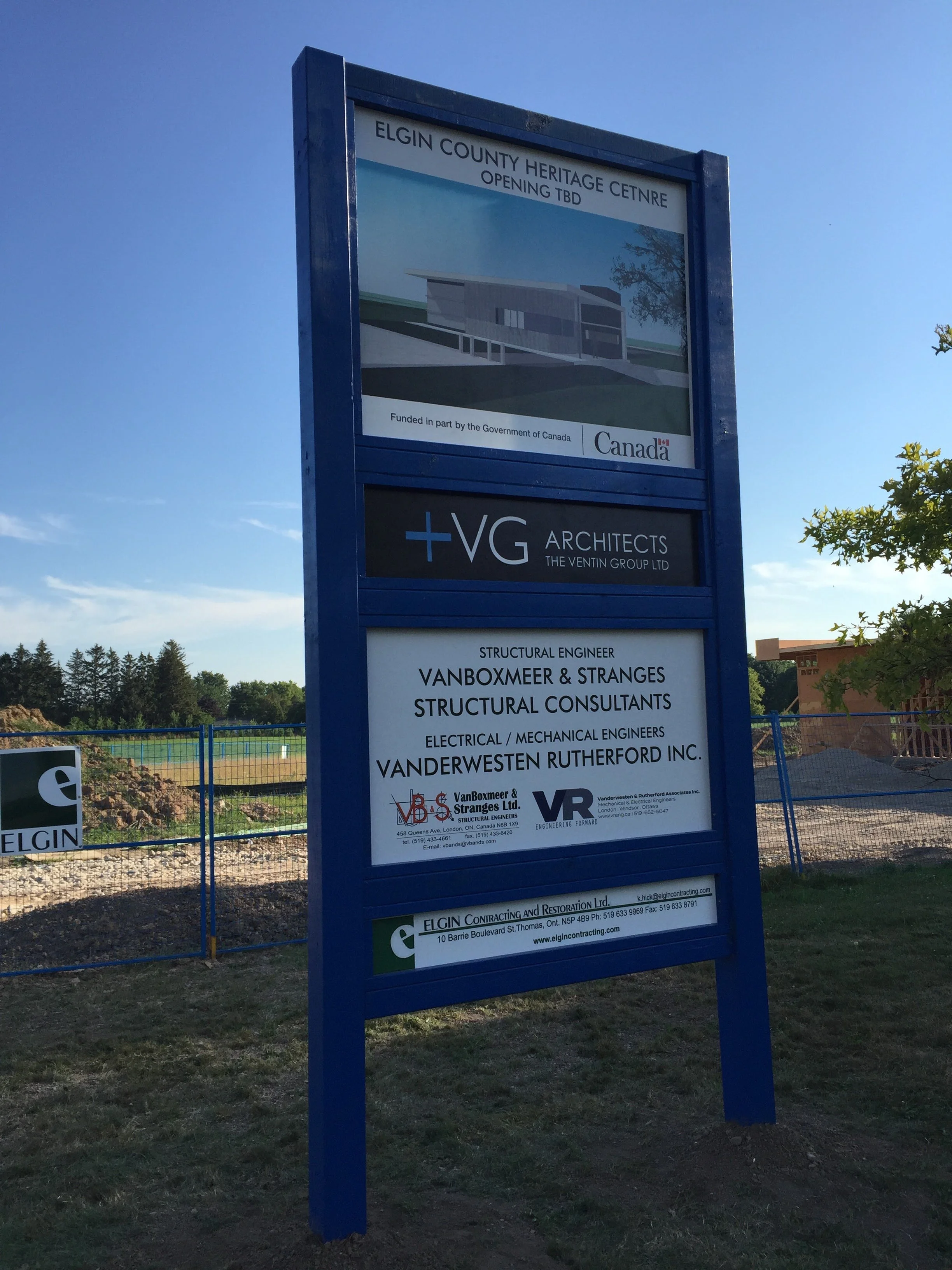 A large blue signboard at a construction site displaying information about the Elgin County Heritage Centre and various architectural and engineering companies involved in the project.