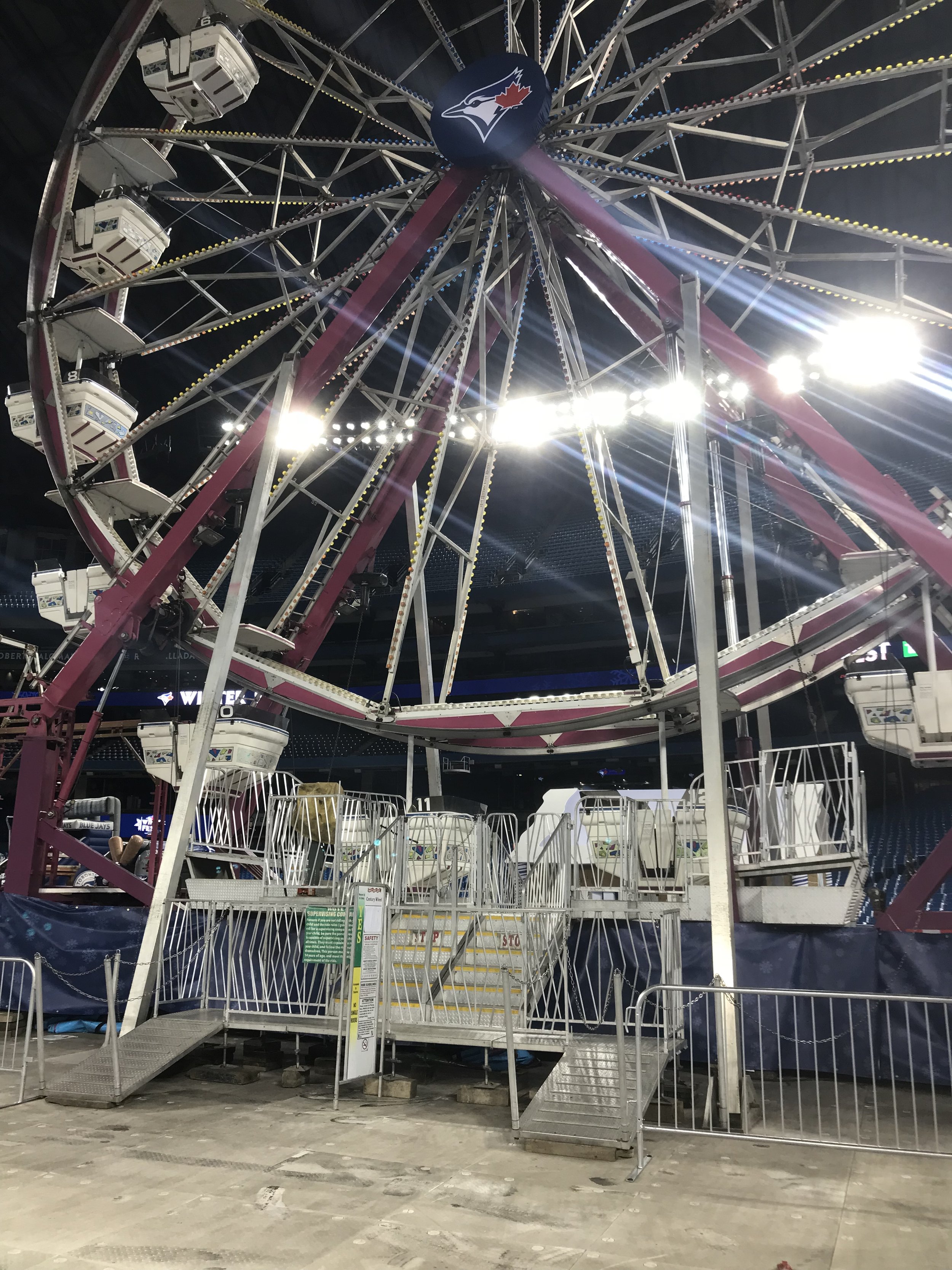 An empty Ferris wheel at a fairground or carnival at night, with bright lights and a Toronto Blue Jays logo at the top.