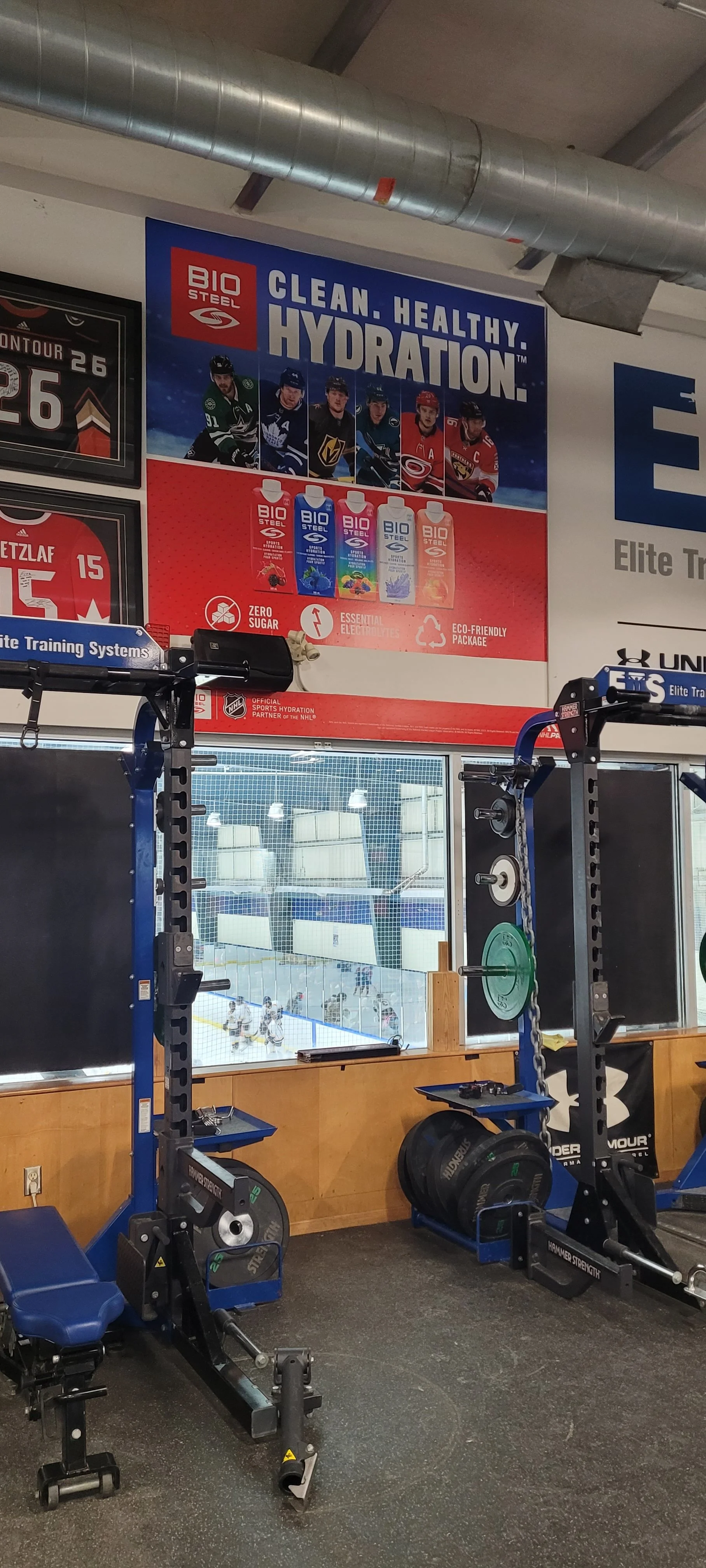 Inside an ice hockey training facility with workout equipment near the windows and a large advertisement poster for Bio Steel hydration drinks above it. The poster features hockey players and highlights hydration benefits.