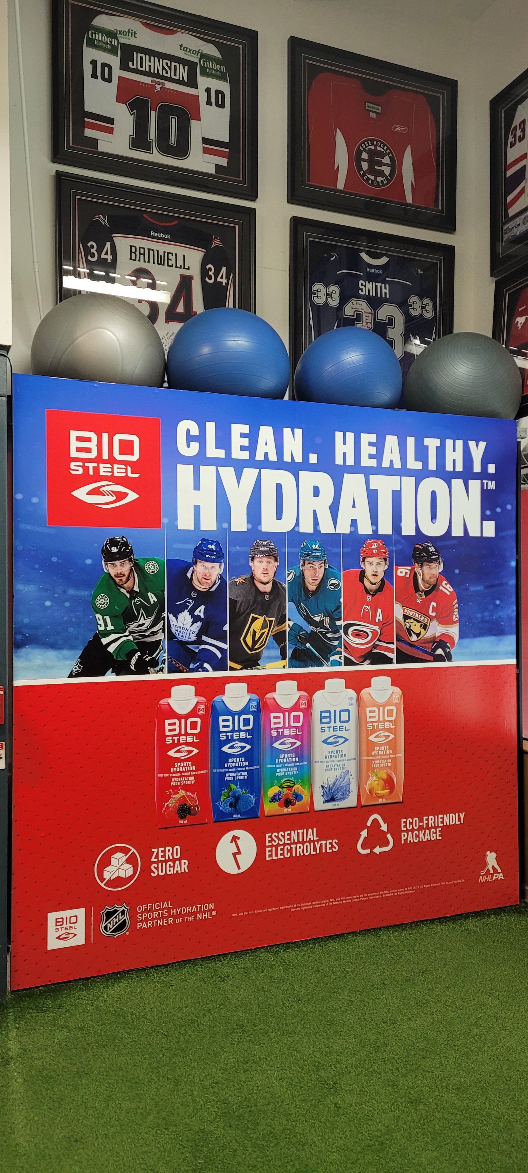 A hockey-themed hydration display featuring a banner advertising Bio Steel sports hydration drinks, emphasizing cleanliness and health. The banner shows images of hockey players and bottles of hydration drinks in various flavors. In the background, there are framed hockey jerseys and equipment, along with exercise balls on top of the display.