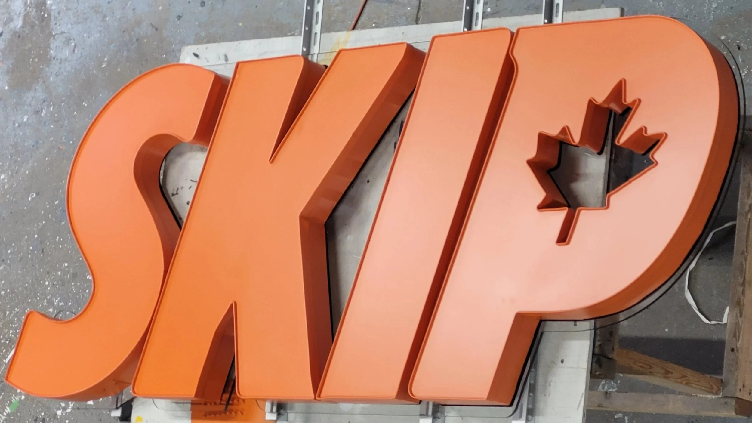 Orange illuminated sign for Skip The Dishes with cutouts of a maple leaf and other shapes, placed on a worktable in a workshop.