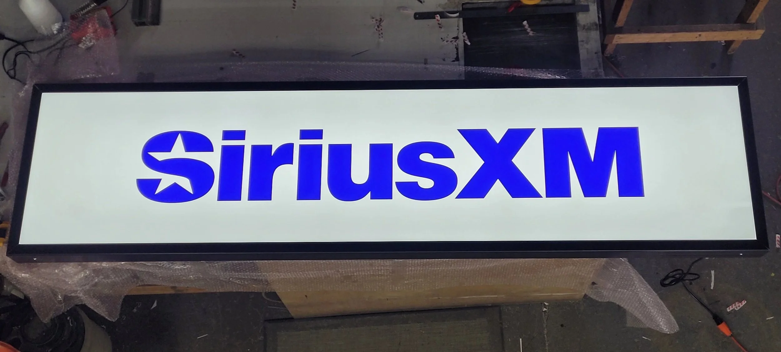 Illuminated sign with the text 'SiriusXM' in blue on a white background, lying flat on a workbench.