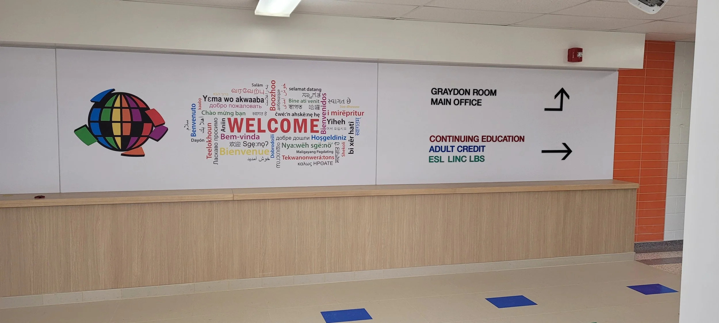 Wall sign with a colorful globe logo and a world of multilingual greetings saying 'Welcome'. The sign contains directions to the 'Graydon Room Main Office' with arrows pointing to the right and upward. It lists languages including English, Spanish, F