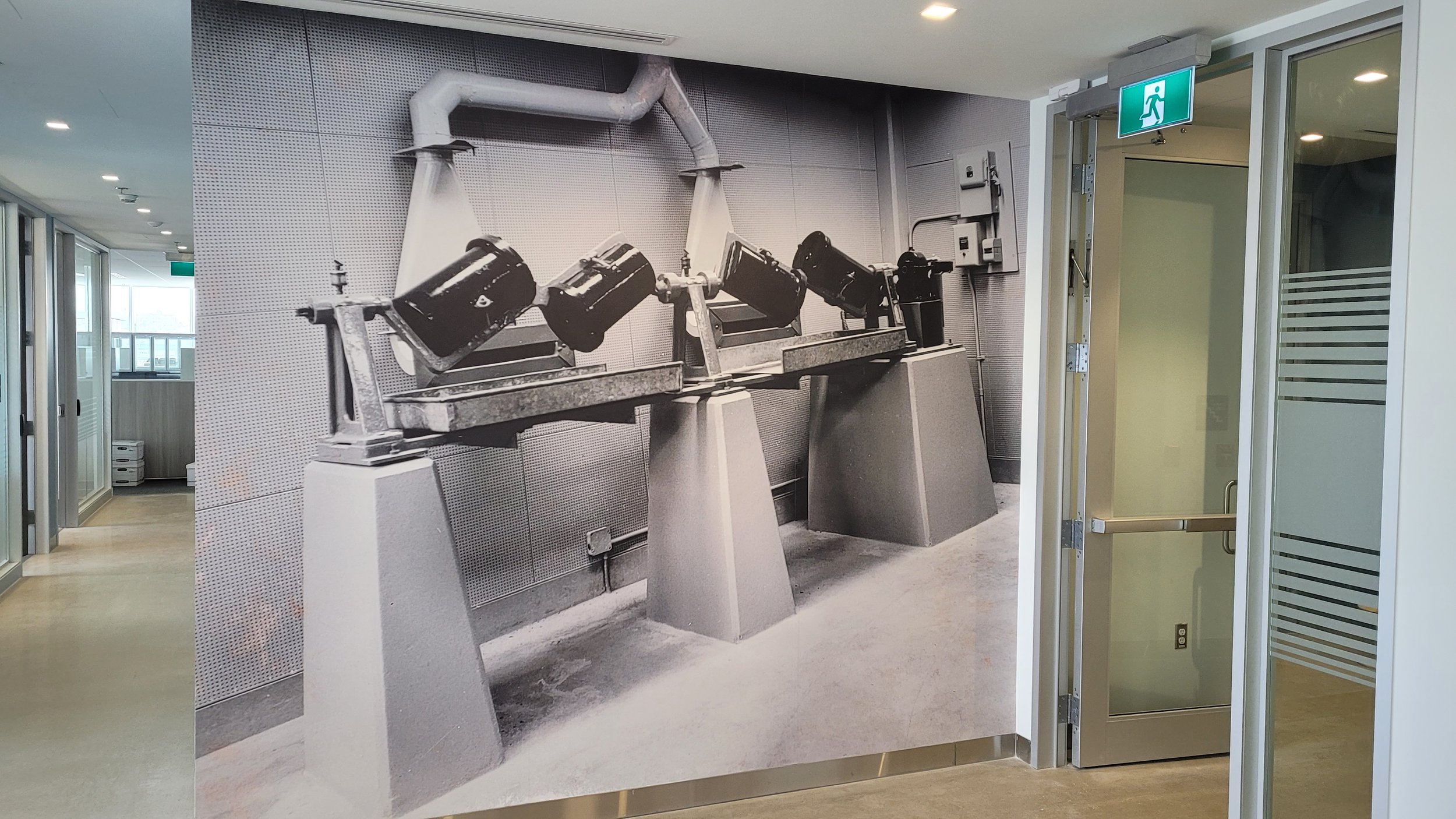 Wall mural of a black-and-white photo showing industrial equipment with pipes and motors, mounted on concrete pedestals inside a modern building corridor.