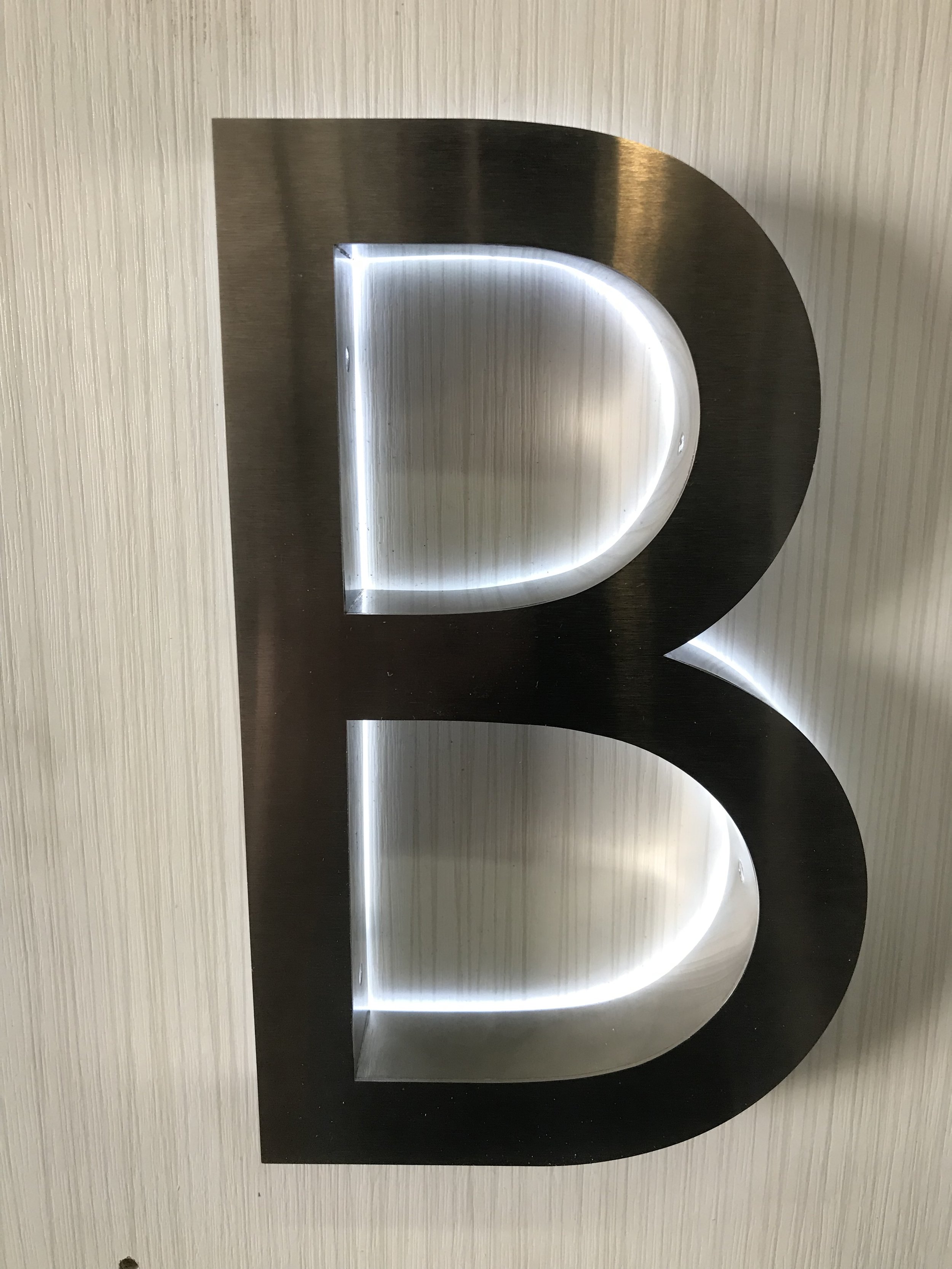 Metal letter B with LED backlighting mounted on a textured wall.