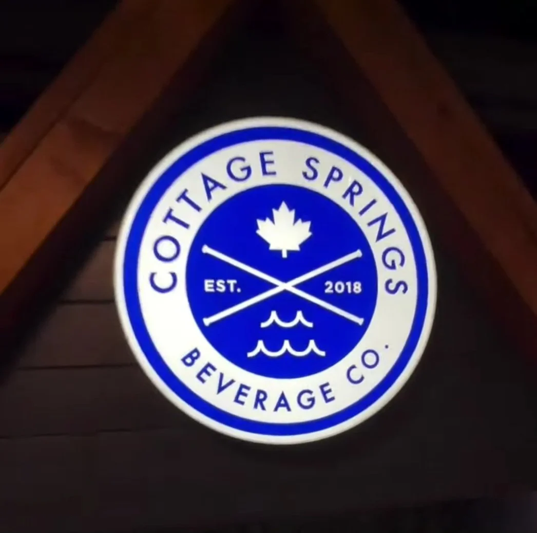 Illuminated circular sign for Cottage Springs Beverage Co., established in 2018, featuring a maple leaf, crossed hockey sticks, and water waves in blue and white.