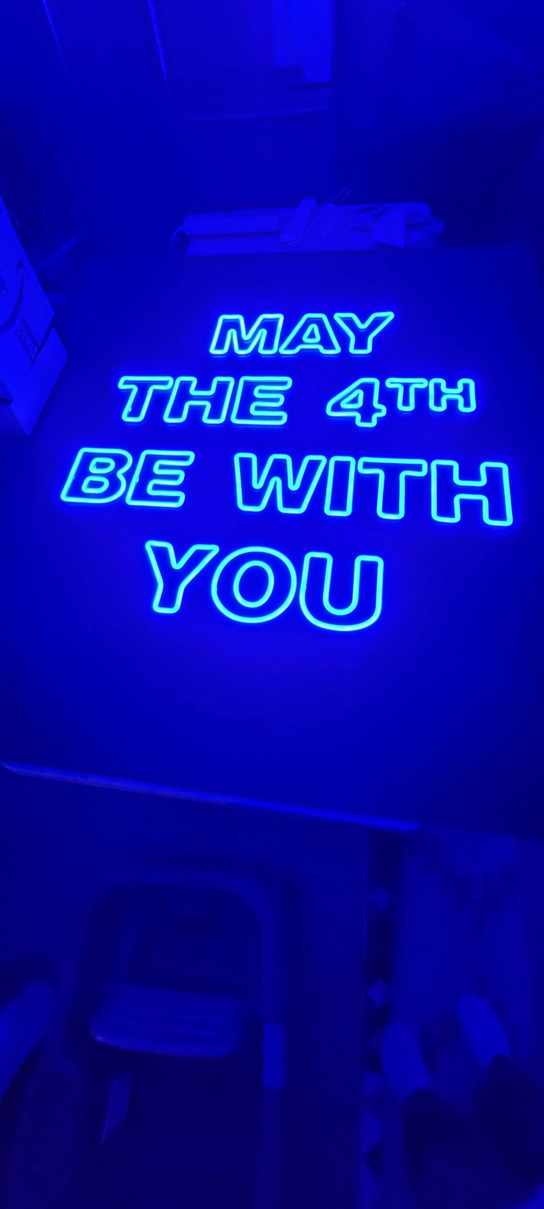 Blue neon sign saying 'May the 4th be with you' on a dark background.
