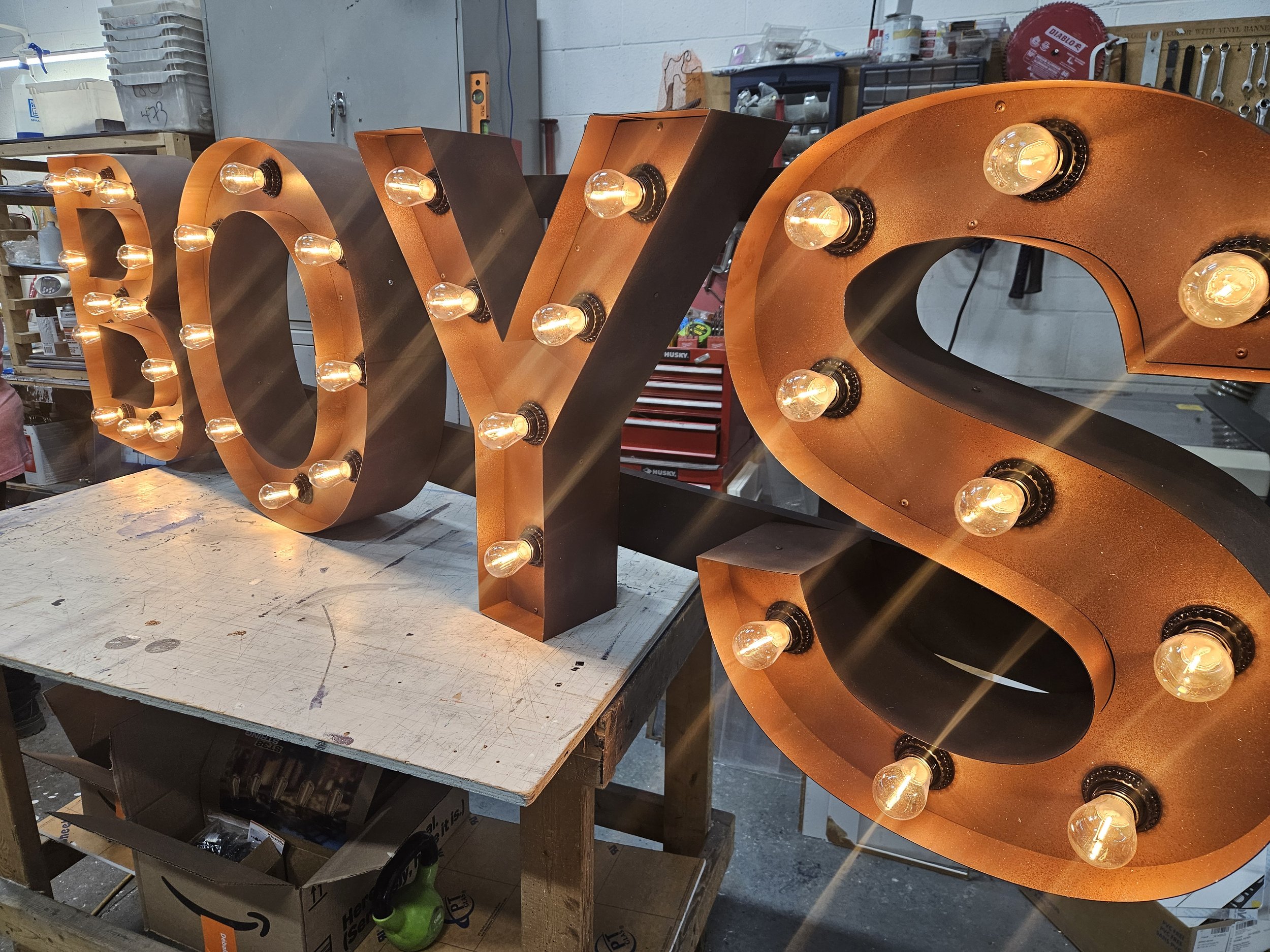 Large decorative marquee letters spelling 'BOYS' with light bulbs inside each letter, placed on a work table inside a workshop.