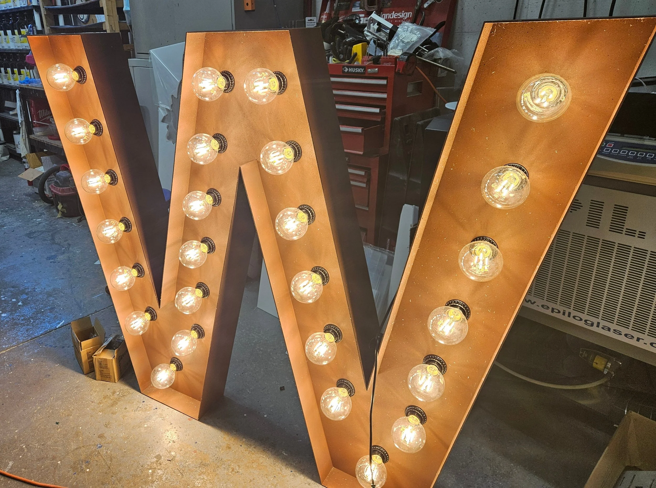 A large decorative letter 'W' illuminated with round light bulbs on a workbench in a workshop. The letter has a wooden front with black edges, and the bulbs are evenly spaced along its length.