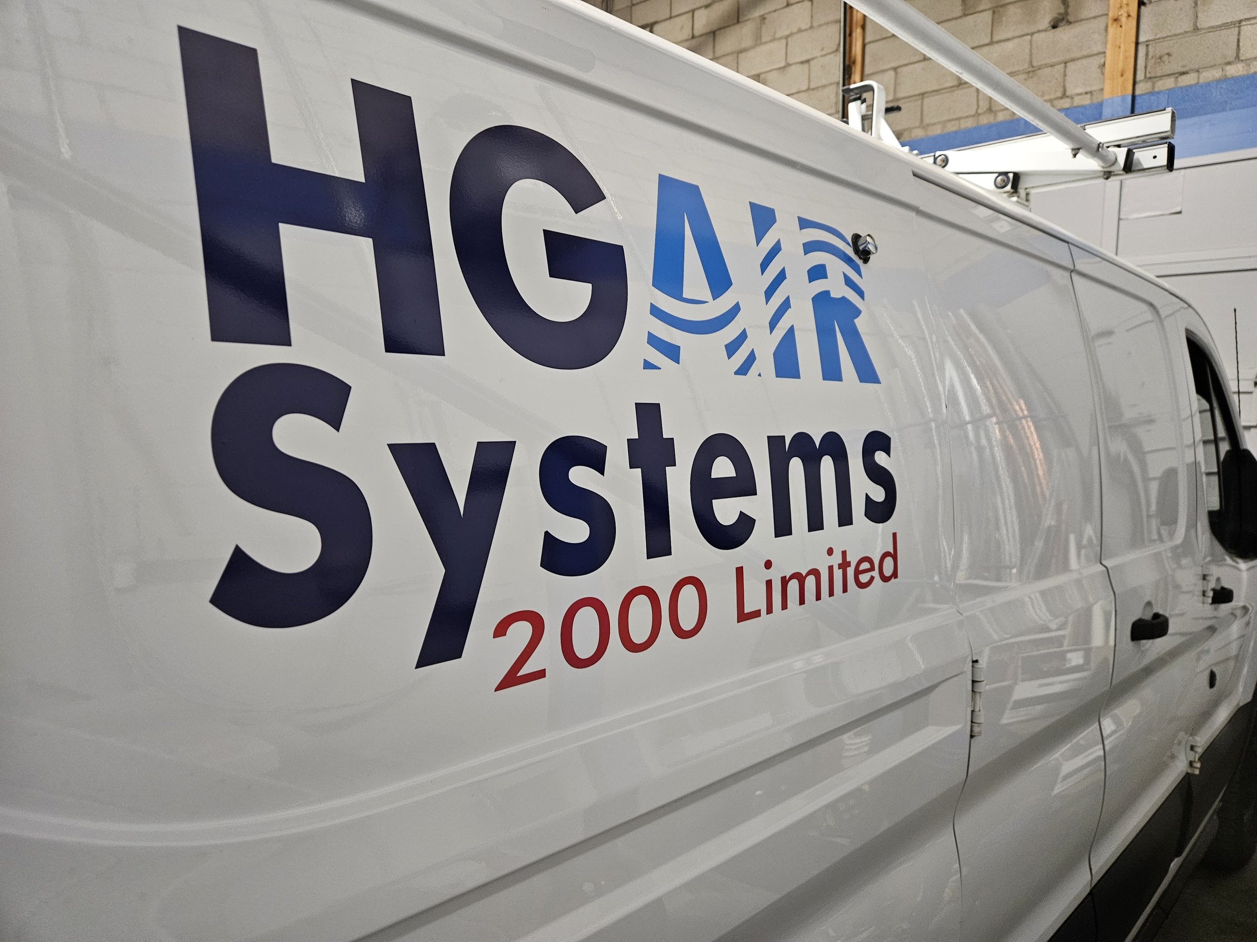 White van with logo reading "HGA Systems 2000 Limited" on side, with blue and red text and design, parked in a warehouse or garage with brick wall and wooden support beams visible.