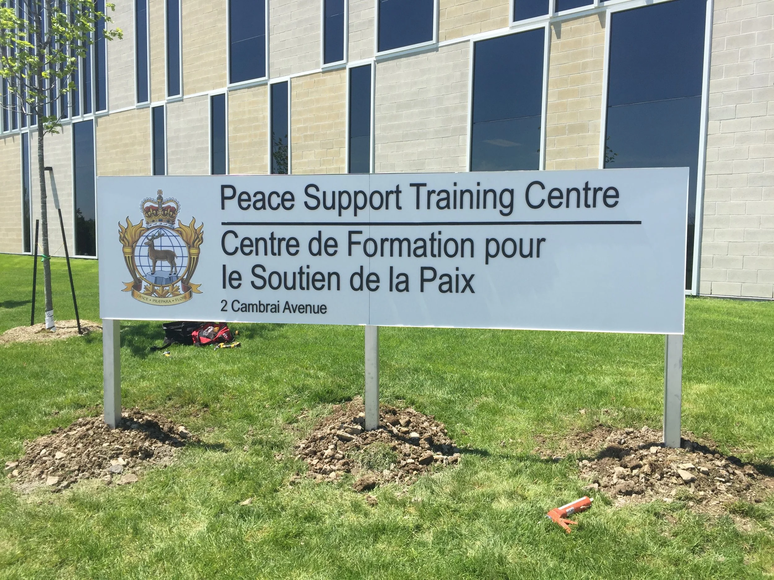 Sign for Peace Support Training Centre with emblem, located at 2 Cambrai Avenue, outside modern building, on a grassy area with trees.