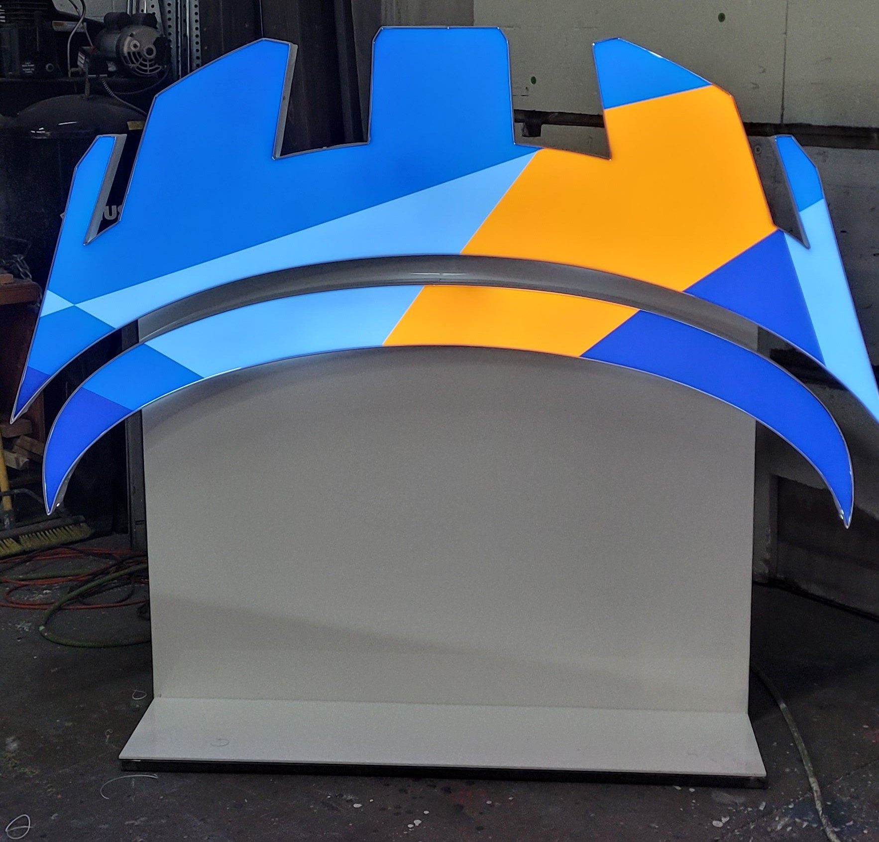 Colorful illuminated sign with geometric shapes in blue, orange, and white, mounted on a gray platform in an industrial setting.