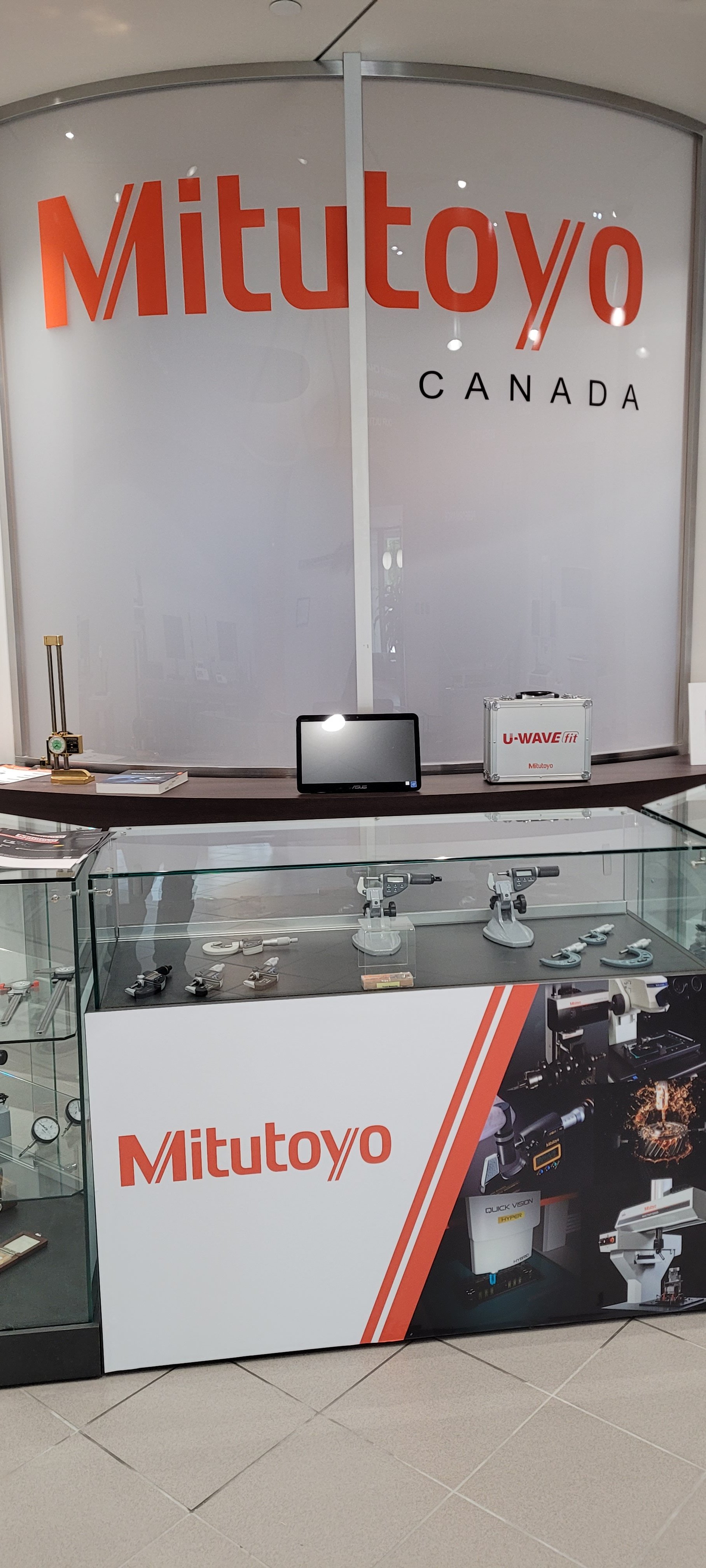 Display case with Mitutoyo tools and equipment, signage, and a counter with various measuring instruments and electronic devices. The background shows a large Mitutoyo Canada logo.