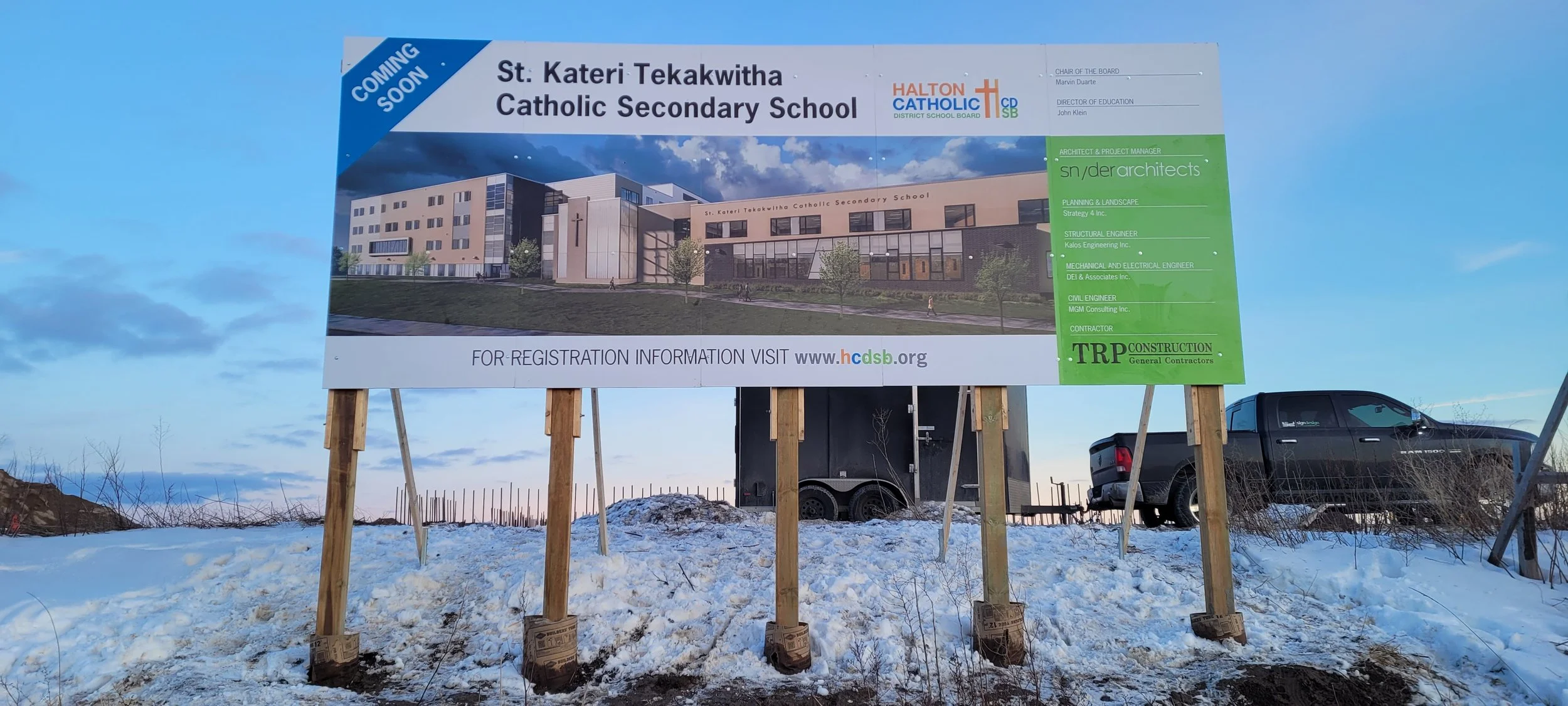 Billboard advertising the construction of St. Kateri Tekakwitha Catholic Secondary School with project details and architect information, set outside in snowy weather with vehicles in the background.