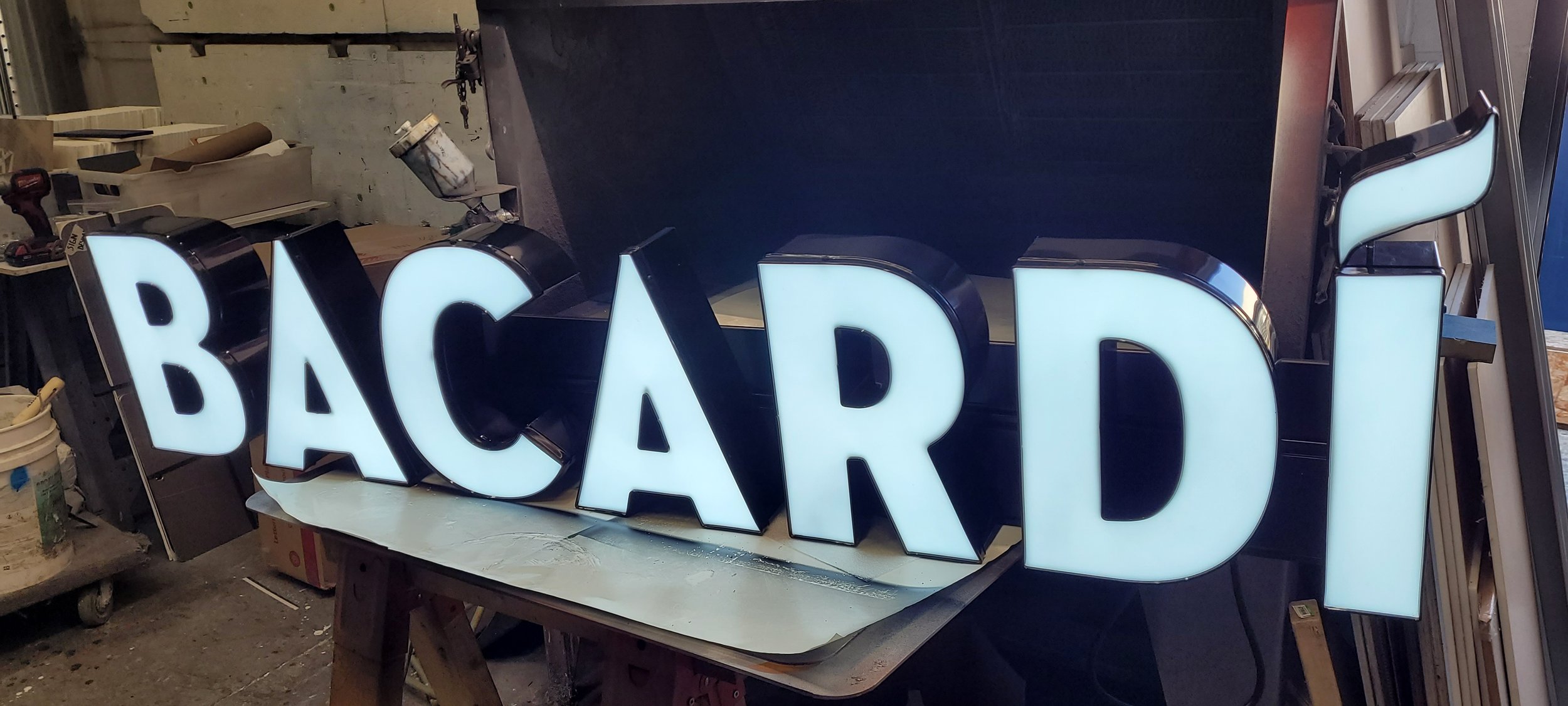 Backlit illuminated sign spelling out the word 'Baccarat' in large white letters with a black border, placed on a wooden workbench in a workshop or storage area.
