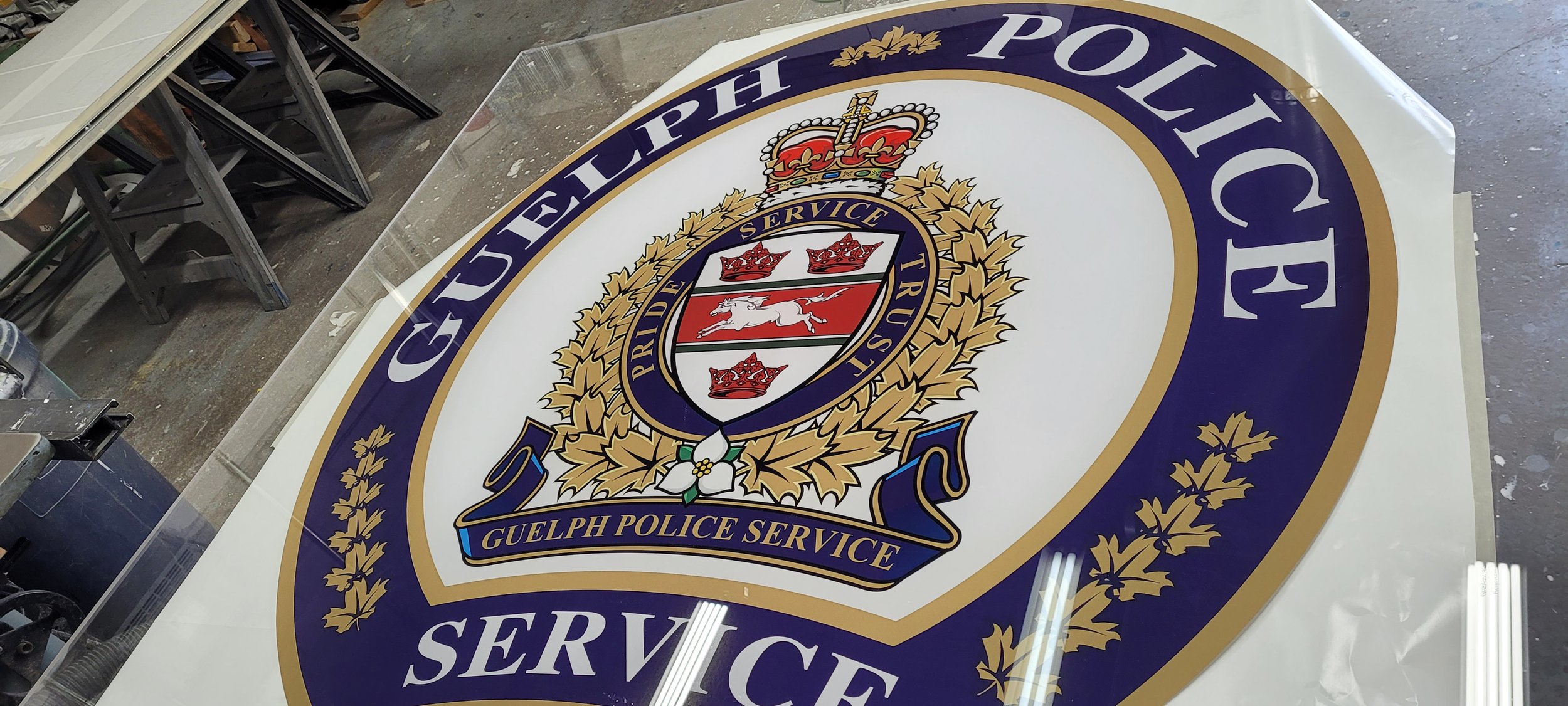 Guelph Police Service badge logo with a blue and gold crown, a white and red shield featuring three crowns, a white cougar, and surrounding gold leaves, with a blue banner at the bottom reading 'Guelph Police Service'.
