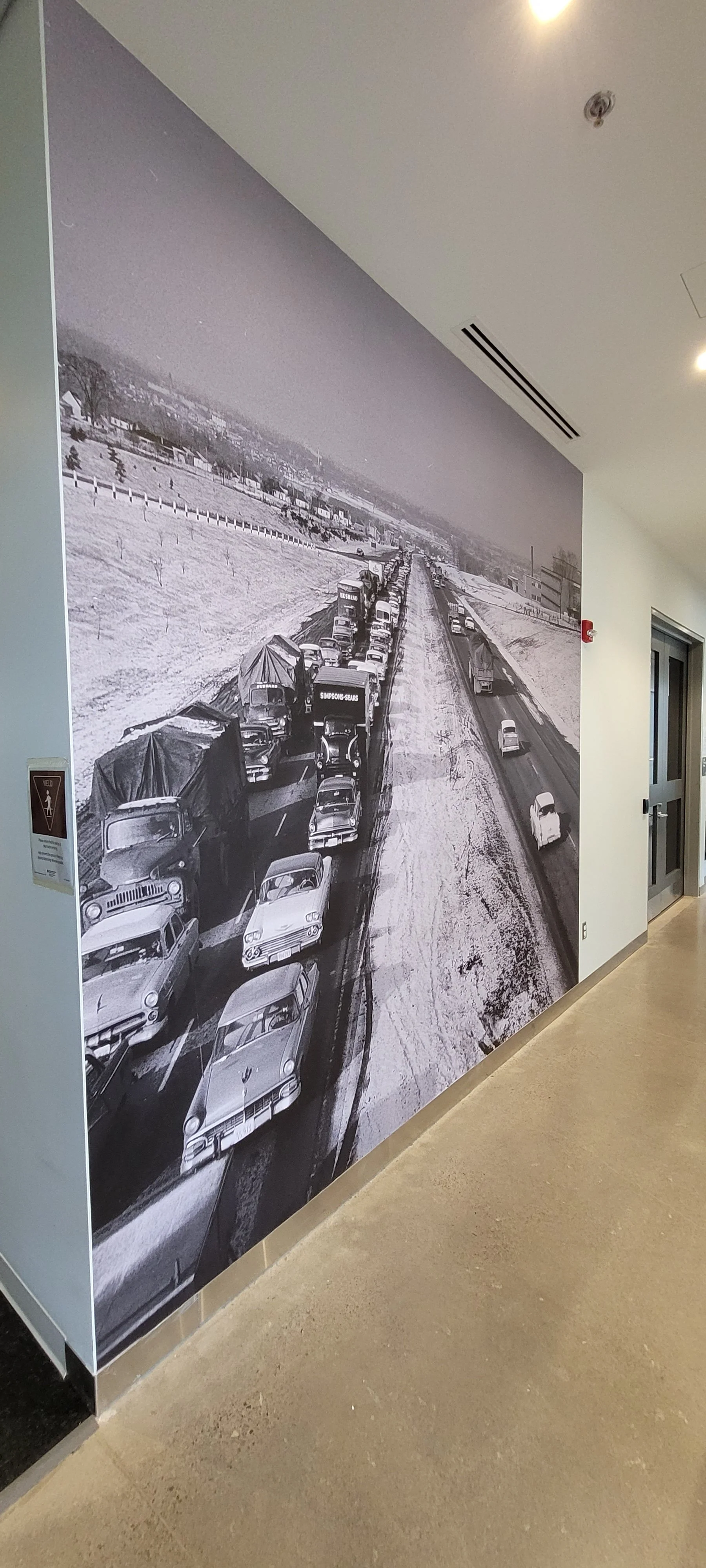 Black and white mural of vintage cars stuck in traffic on a snowy road, with a landscape of open fields and houses in the distance.