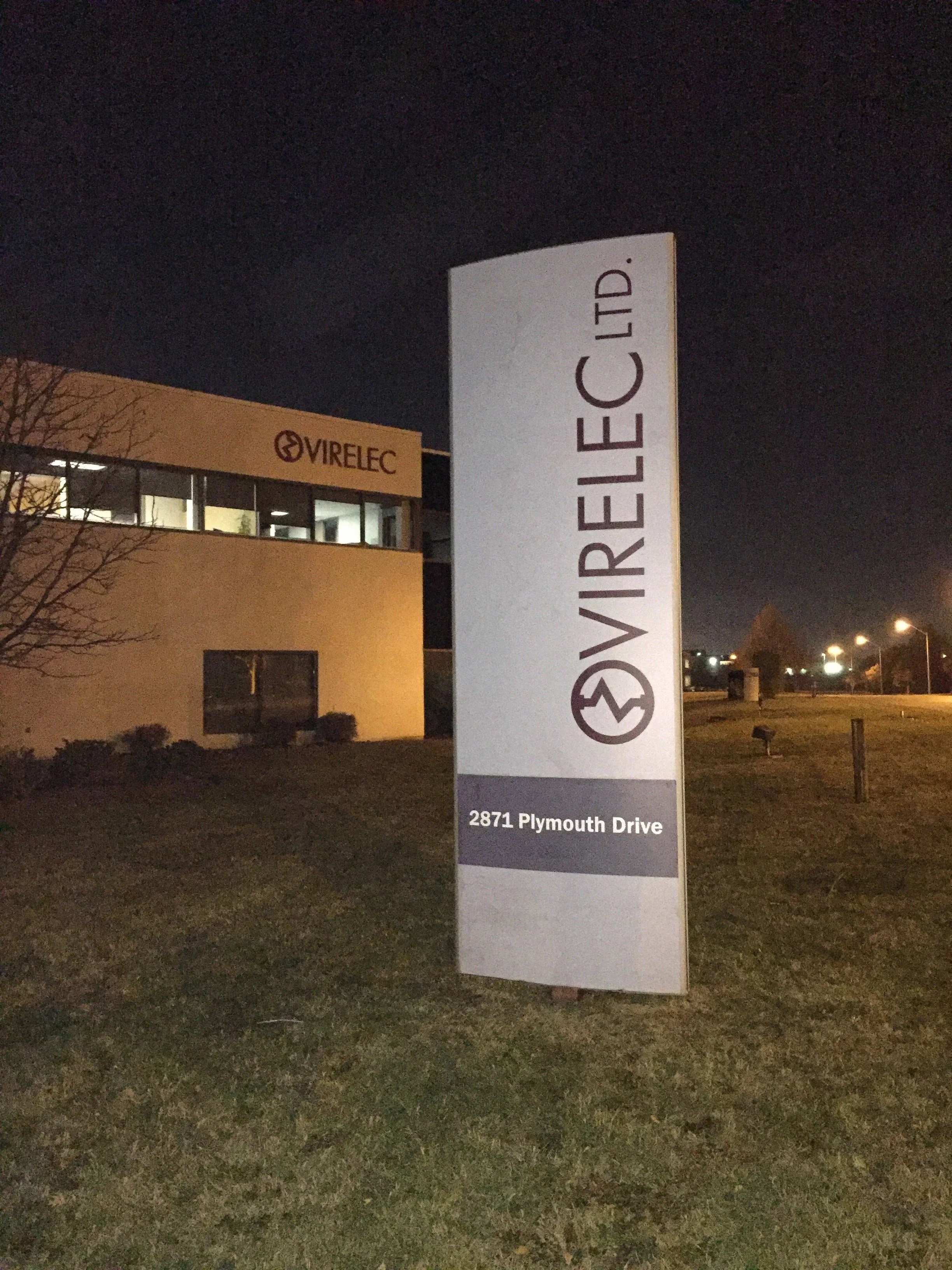 A tall signboard displays the name 'VIRELEC LTD.' with a logo, and the address '2871 Plymouth Drive' at night, with a building in the background that has another 'VIRELEC' sign.