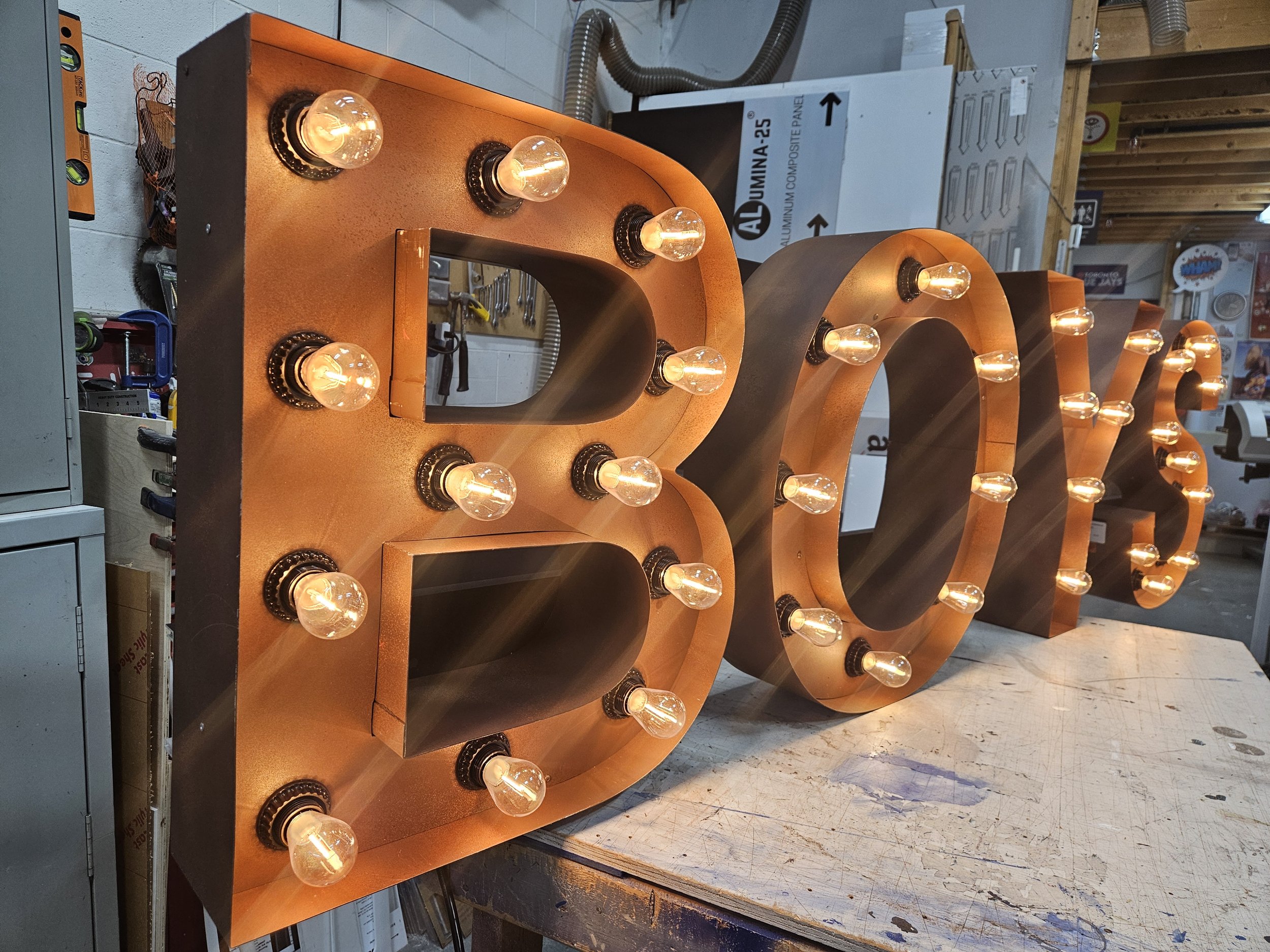 A large decorative sign spelling 'BOYS' with illuminated light bulbs around each letter, resting on a workbench in a workshop.