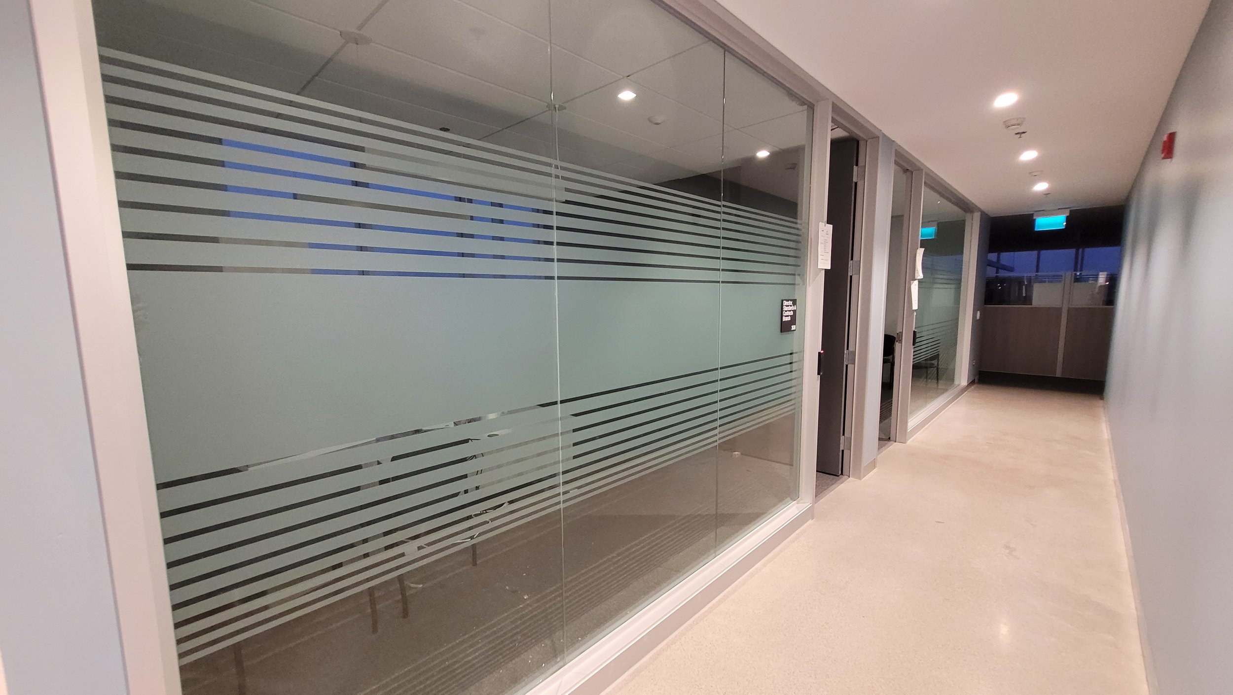 Empty hallway with glass-walled offices and frosted glass with horizontal stripes, during evening or night, with interior lighting and exit signs.