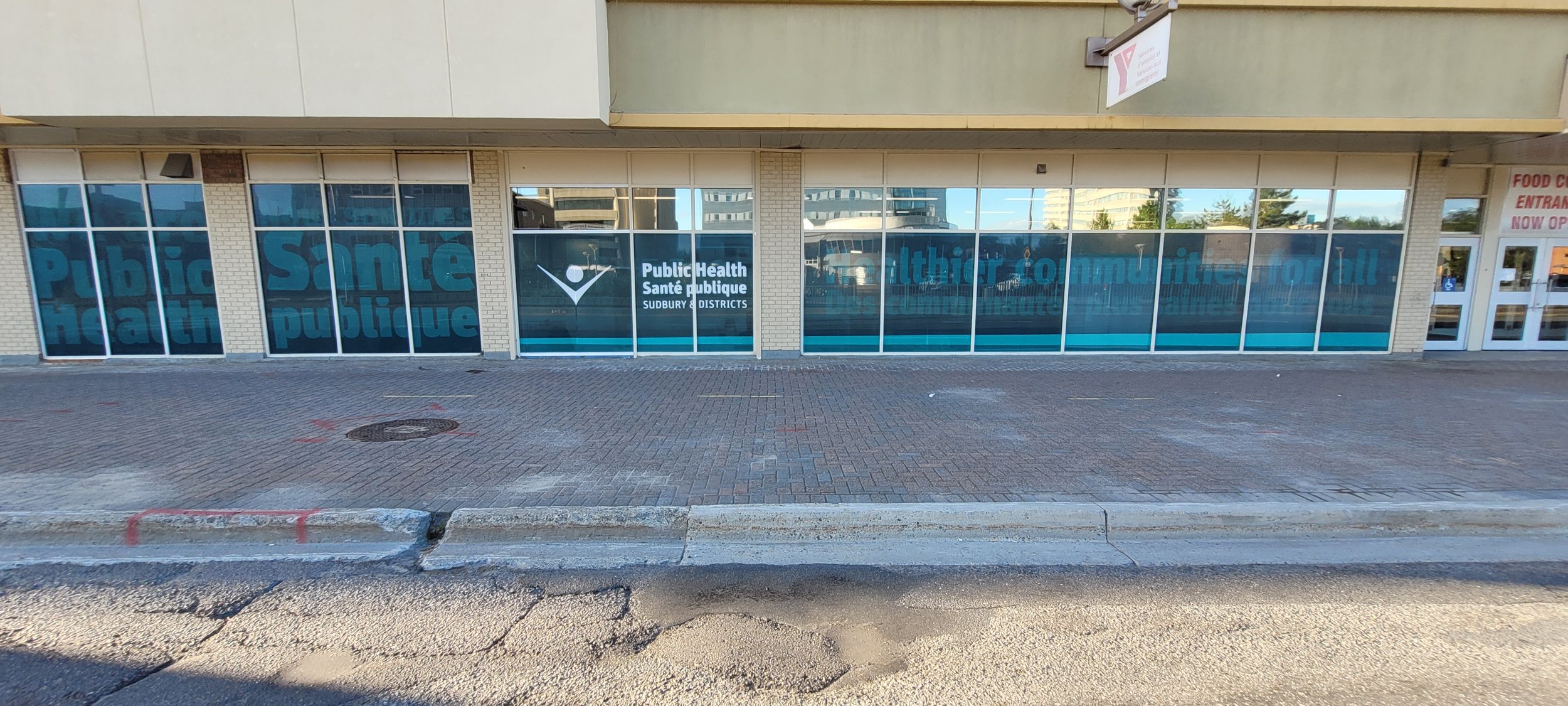 Exterior of a public health clinic with large windows displaying the words 'Public Health' repeatedly. The clinic is located in an urban area with a brick sidewalk in front and a cracked curb in the foreground. There is a sign on the building that re