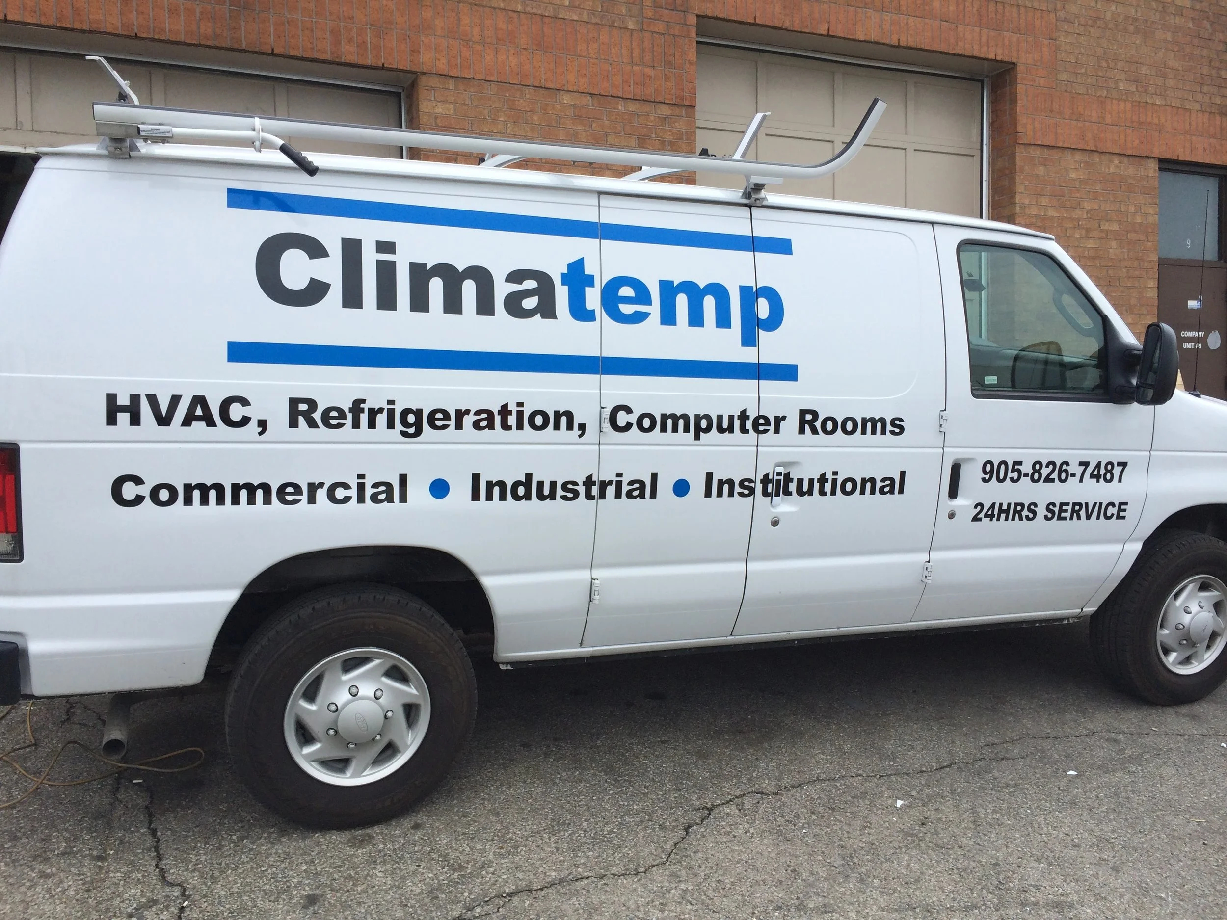 White service van with blue and black lettering for Climate temp, specializing in HVAC, refrigeration, and computer rooms, parked on a paved street in front of a brick building, with a ladder mounted on top and a phone number and 24-hours service wri