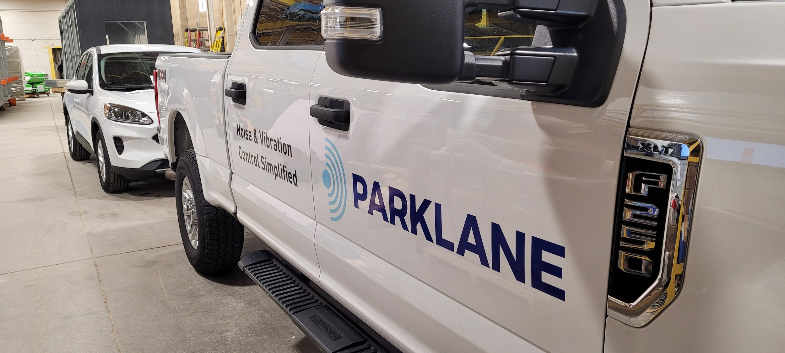 White service truck with 'Parklane' branding and claims of 'Noise & Vibration Control Simplified', parked inside warehouse next to a white sedan vehicle.
