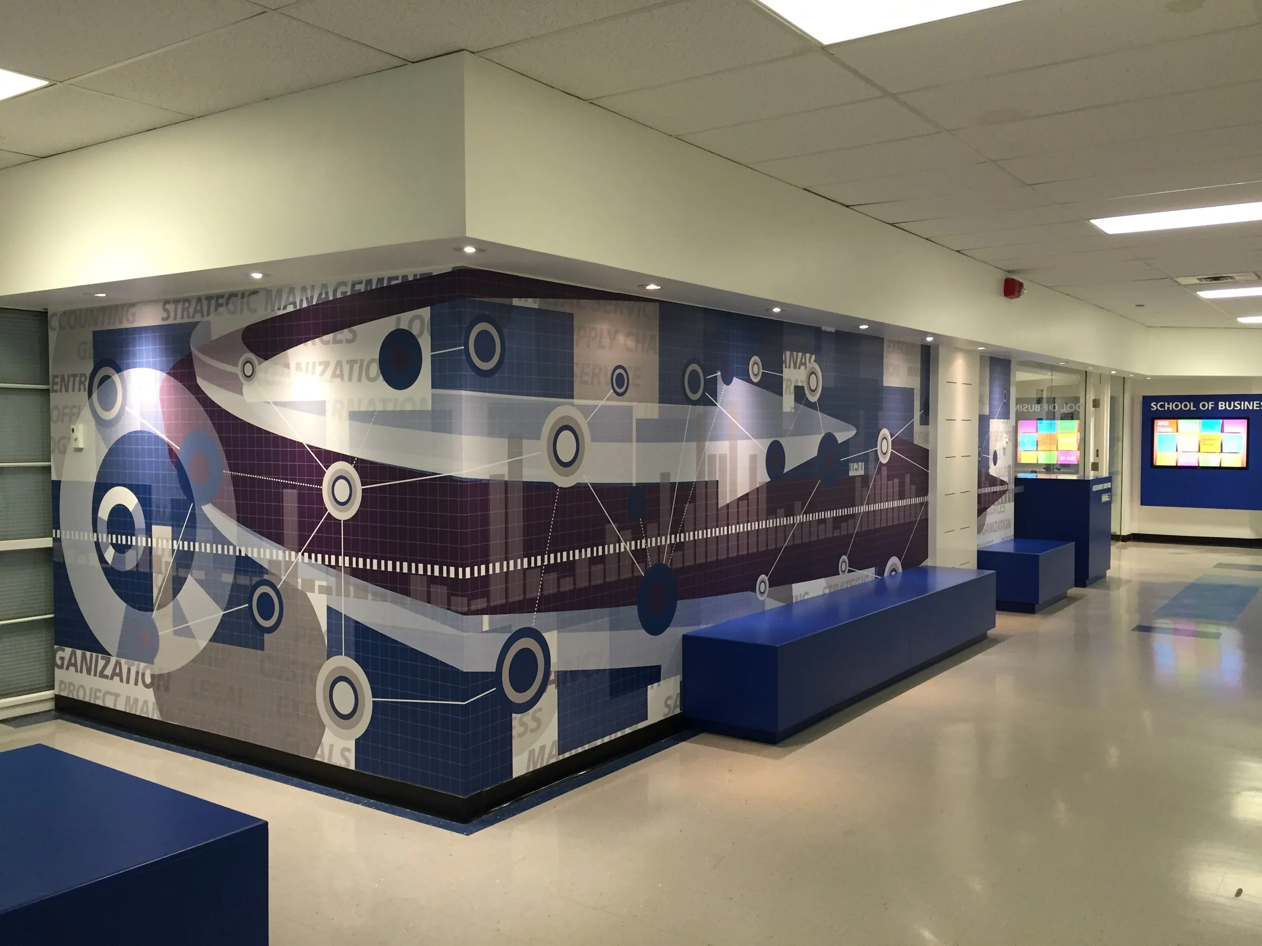 Interior view of a school corridor with a large wall mural depicting interconnected circles and lines, and words like 'Strategic Management,' 'Organization,' and 'Project Management.' There are blue benches along the wall and a blue bulletin board to