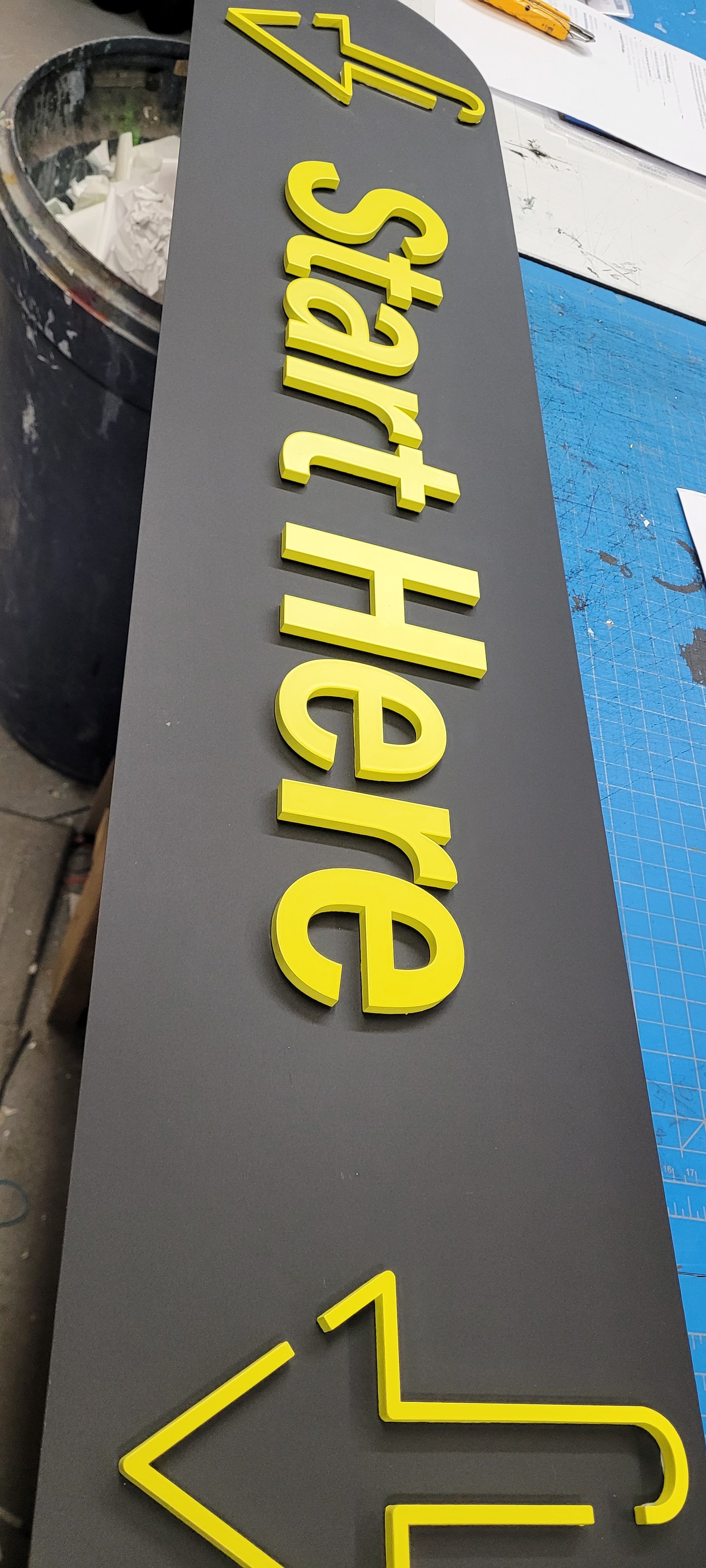 A gray sign with yellow 3D letters that say "Start Here" and an arrow pointing left. The sign is on a workspace with papers and a blue cutting mat visible.
