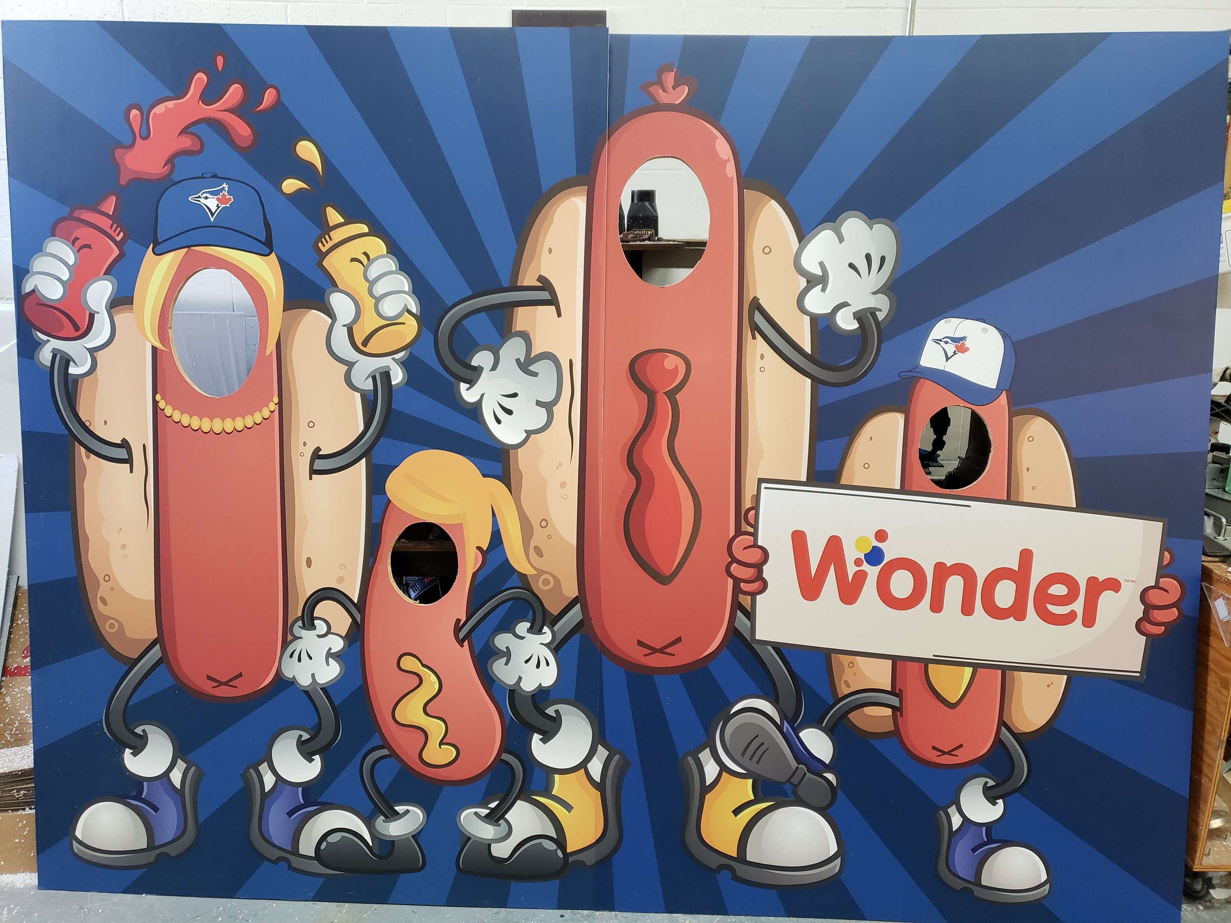 Cartoon characters dressed as hot dogs with arms and legs, some wearing Toronto Blue Jays caps, holding ketchup and mustard bottles, and a sign that says 'Wonder'.