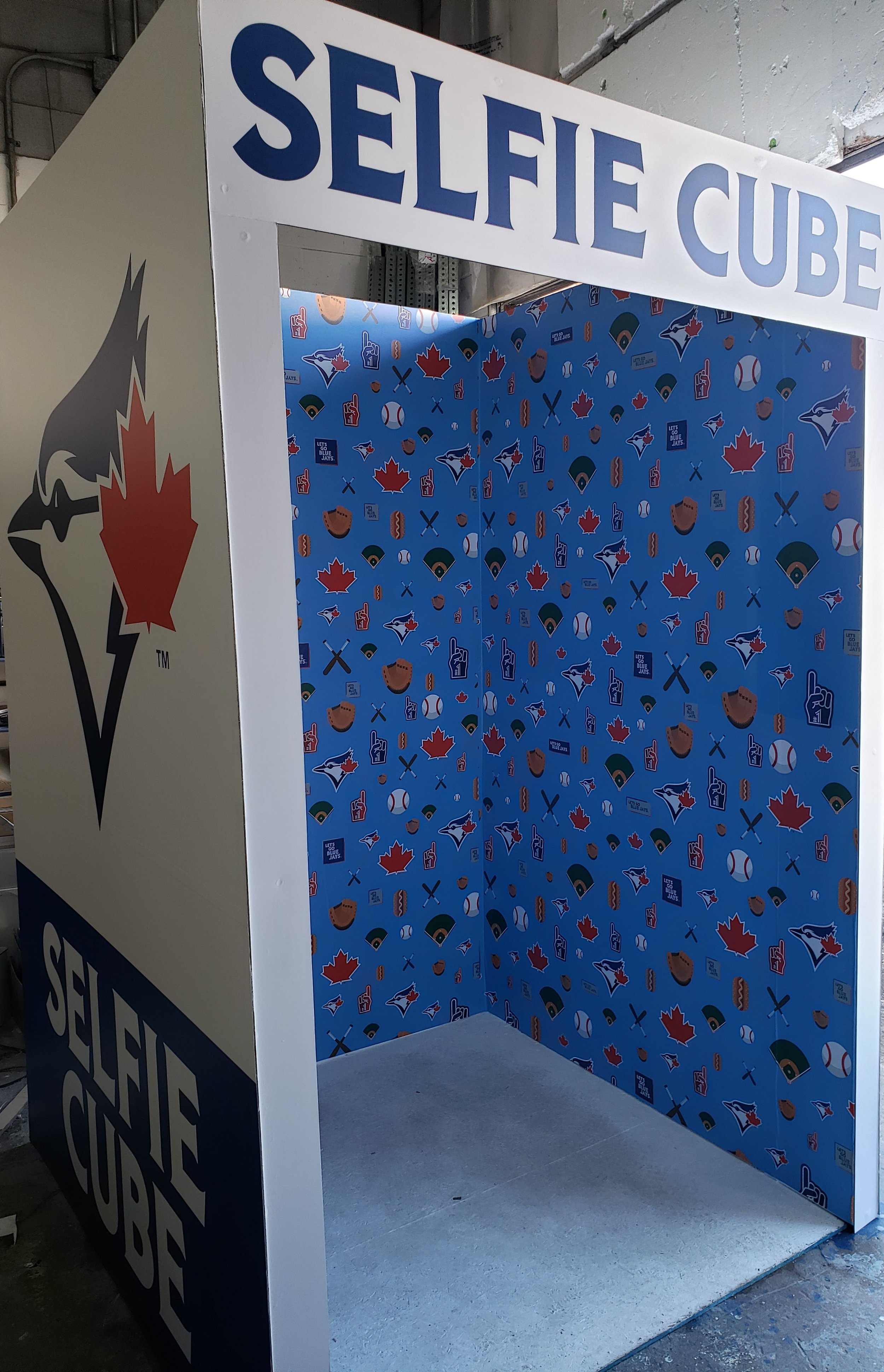 A selfie cube photo booth with blue interior walls featuring Toronto Blue Jays sports team logos and related icons, and white exterior panels with a black and red maple leaf logo and large blue text reading 'SELFIE CUBE' at the top.