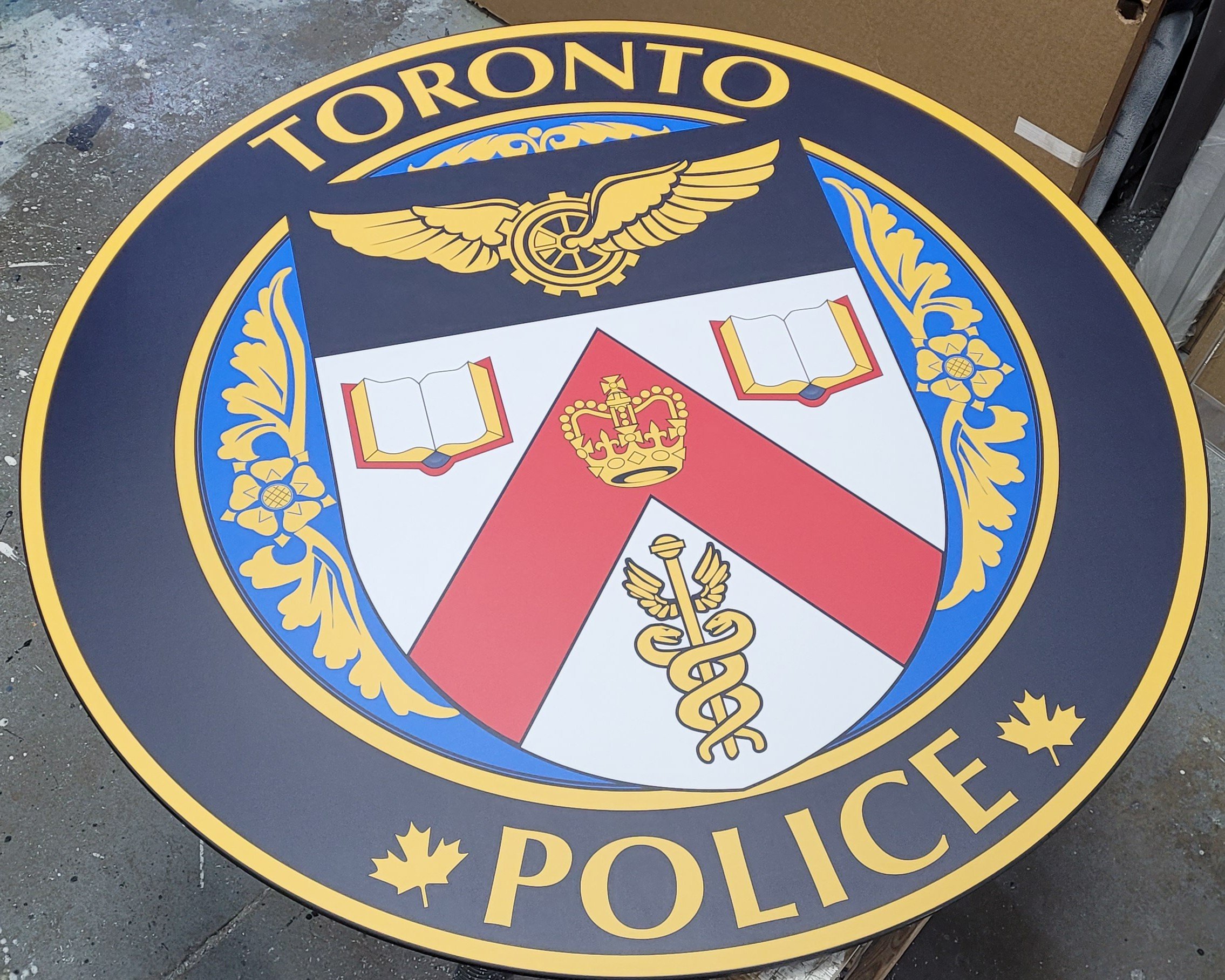 Round Toronto Police emblem with a shield featuring a crown, open books, a caduceus, and wings, surrounded by gold and blue decorations, and the words 'Toronto Police' at the top and bottom.