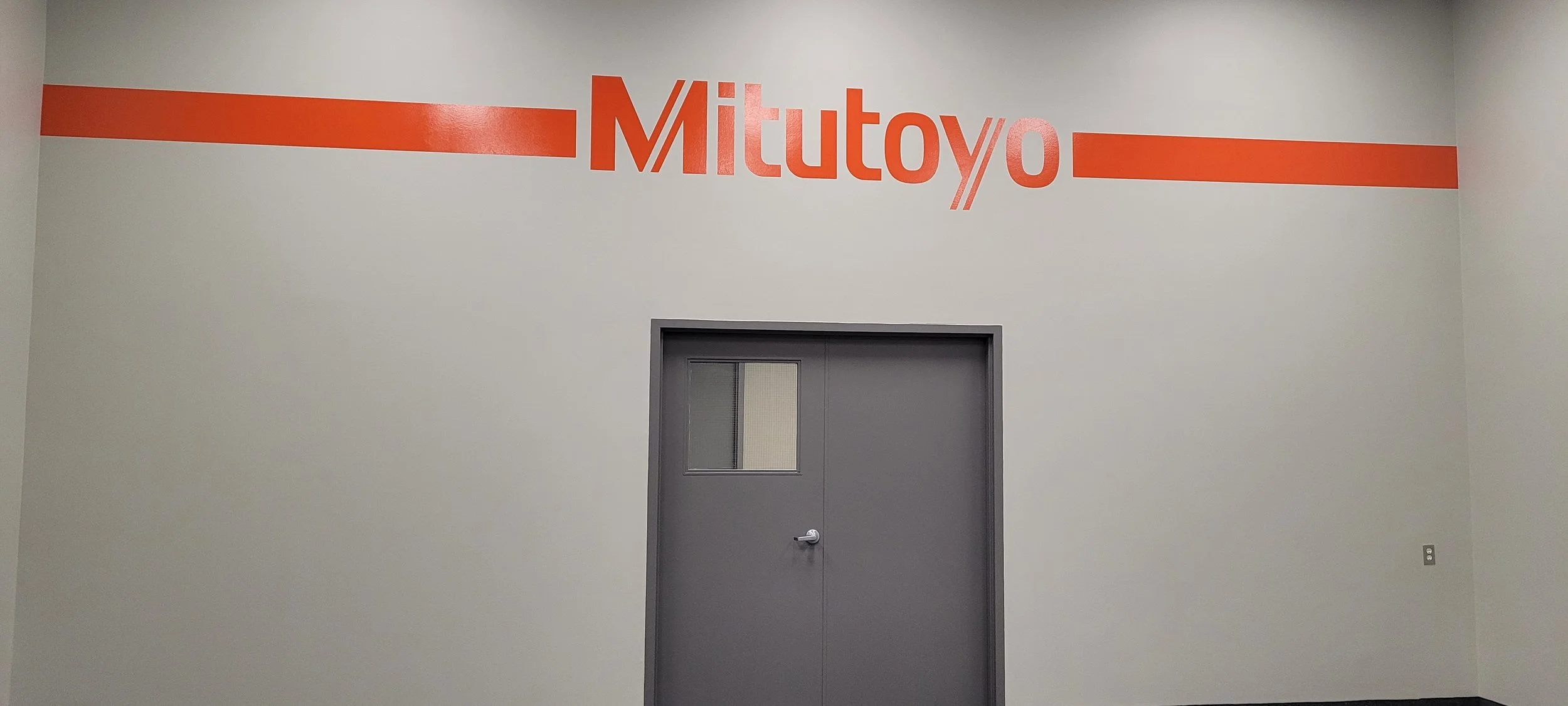 Indoor wall with red and orange coloring, displaying the word 'Mitutoyo' in large letters above a gray door with a small window.