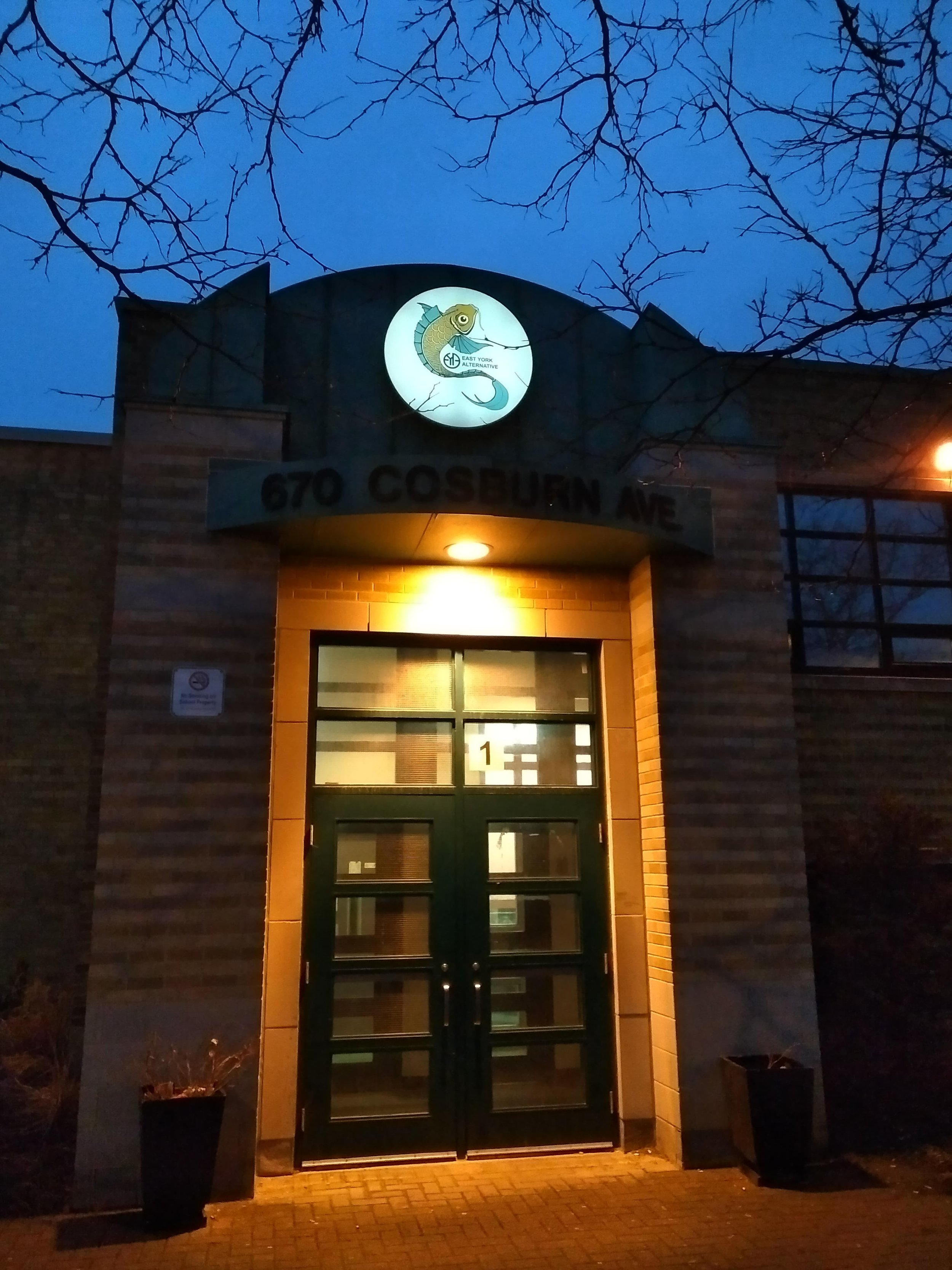 Nighttime view of a brick building entrance with double glass doors, illuminated by a wall-mounted light above. An illuminated round sign features a stylized fish with the text 'East York Alternative.' Two potted plants are placed on either side of t