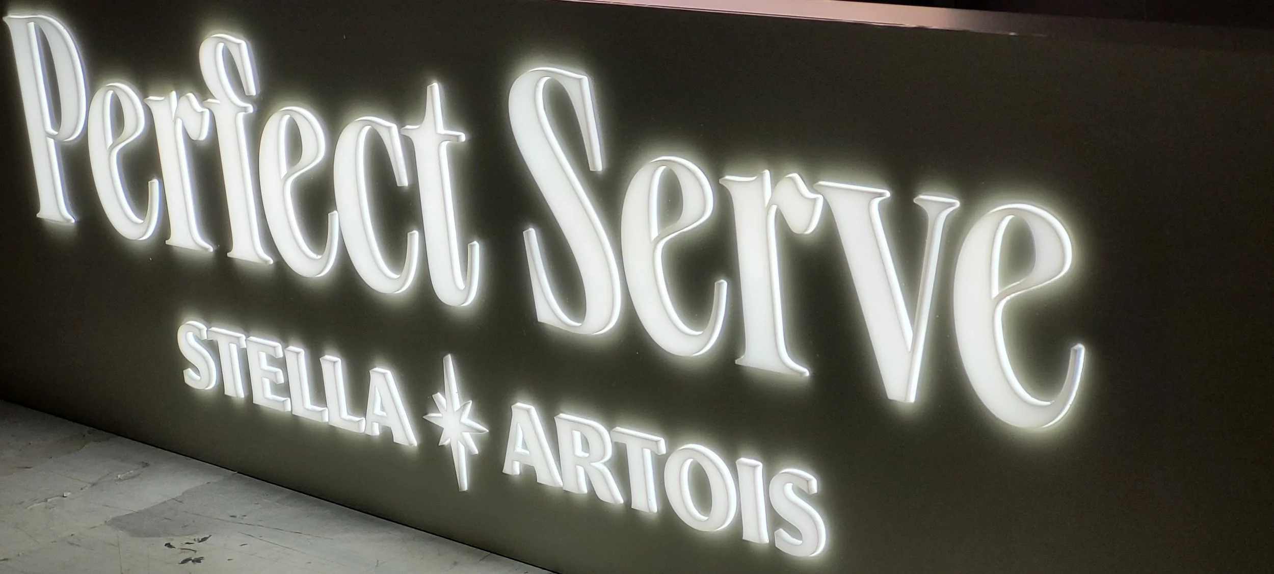 Neon sign with the words 'Perfect Serve' in cursive font and 'Stella Artois' in uppercase letters, with a star symbol between 'Stella' and 'Artois'.