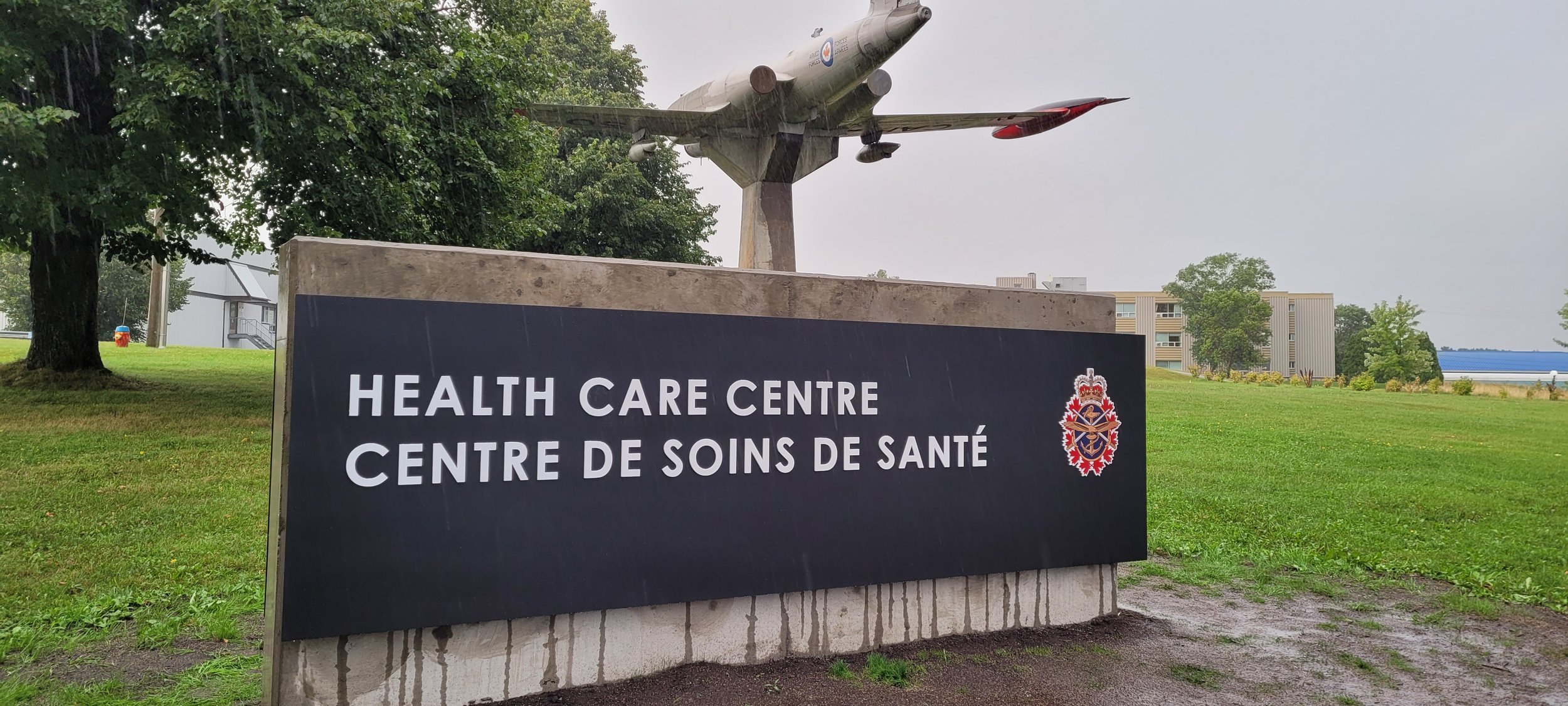 Sign for a healthcare centre with a military aircraft monument on top, situated outdoors amidst greenery and buildings.