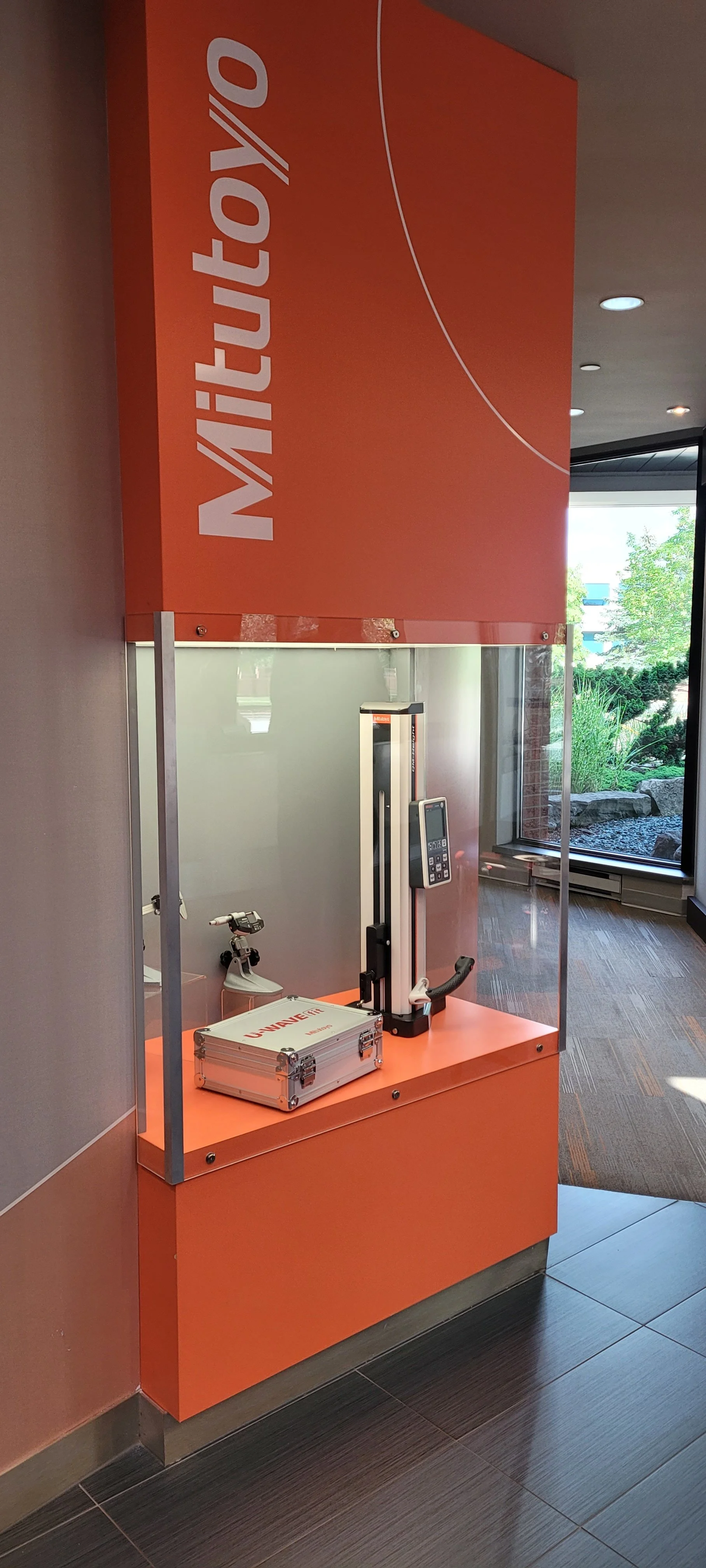 A vertically oriented display case with an orange background and white text that reads 'METUTUL'O'. Inside the case, there is a height measurement device, a tablet, and a small machine labeled 'UNIQUE FIT'. The display is located near a window with a view of trees and rocks outside.