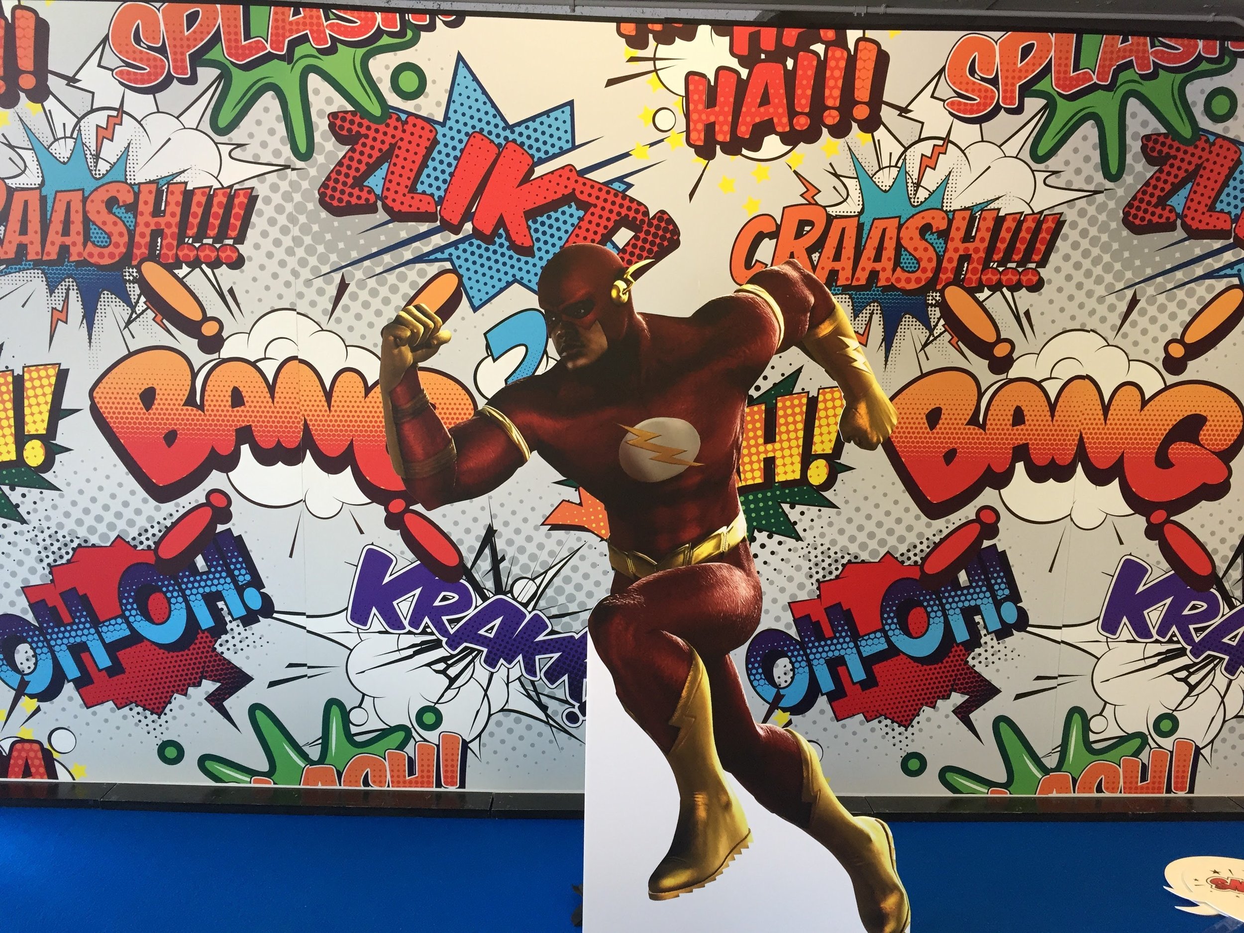 The image features a superhero mascot in a costume with a lightning bolt emblem on the chest, striking a dynamic pose with one arm flexed and the other bent. The background is decorated with vibrant comic book style words like `BAM!`, `CRASH!`, `KA-B