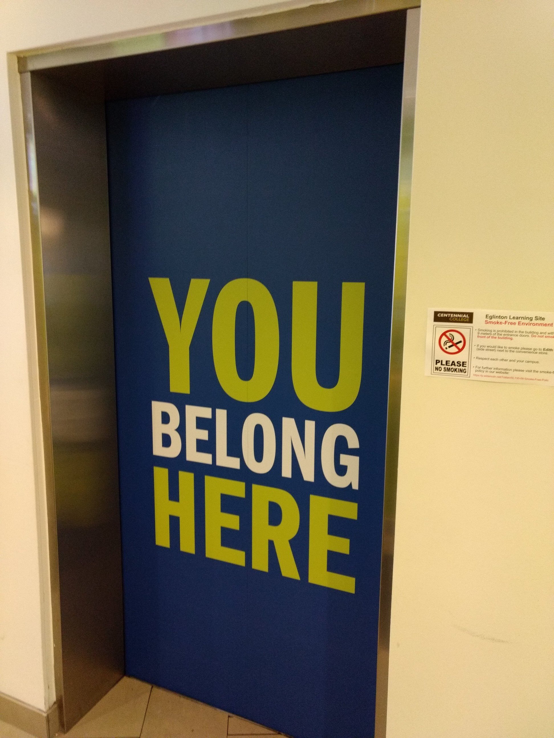 Blue elevator door with the words "YOU BELONG HERE" written on it in large yellow and white letters.