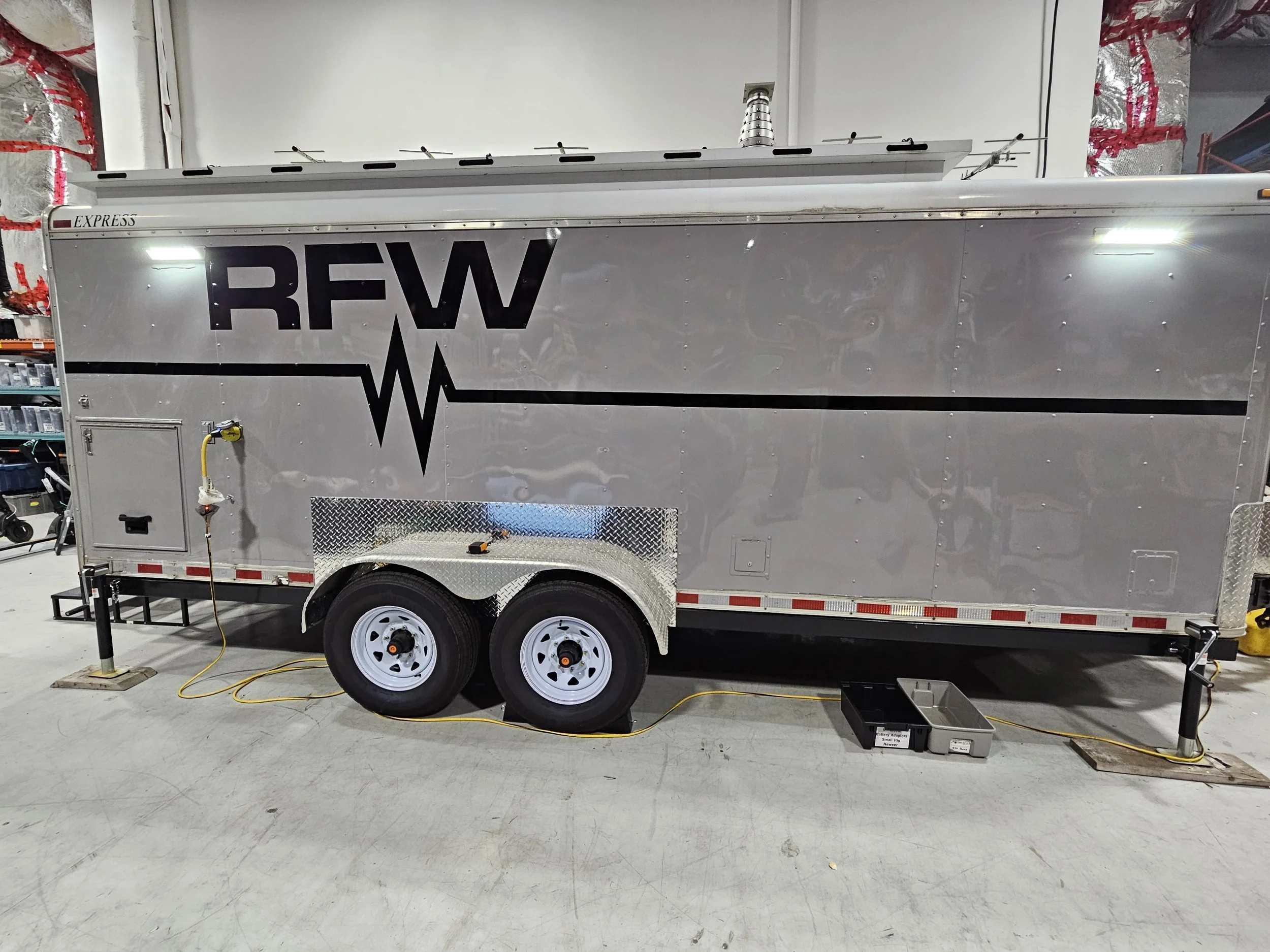 A silver mobile medical trailer with the logo 'RFW' and a heartbeat line graphic, parked indoors on a concrete floor, connected to power.
