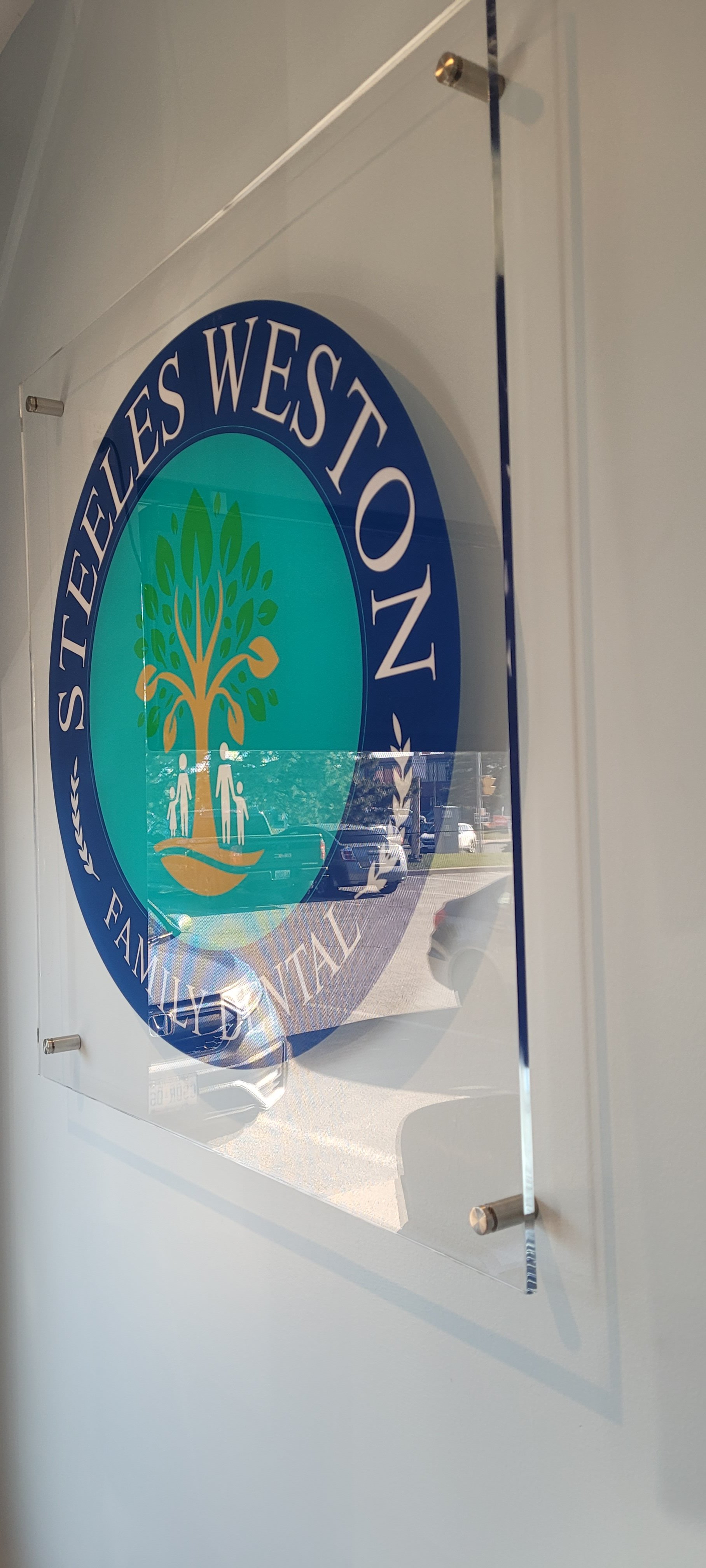 A sign on a wall for the Tyees Westown Family Dental office, featuring a logo with a tree and family figures, and an outside view reflected in the glass.