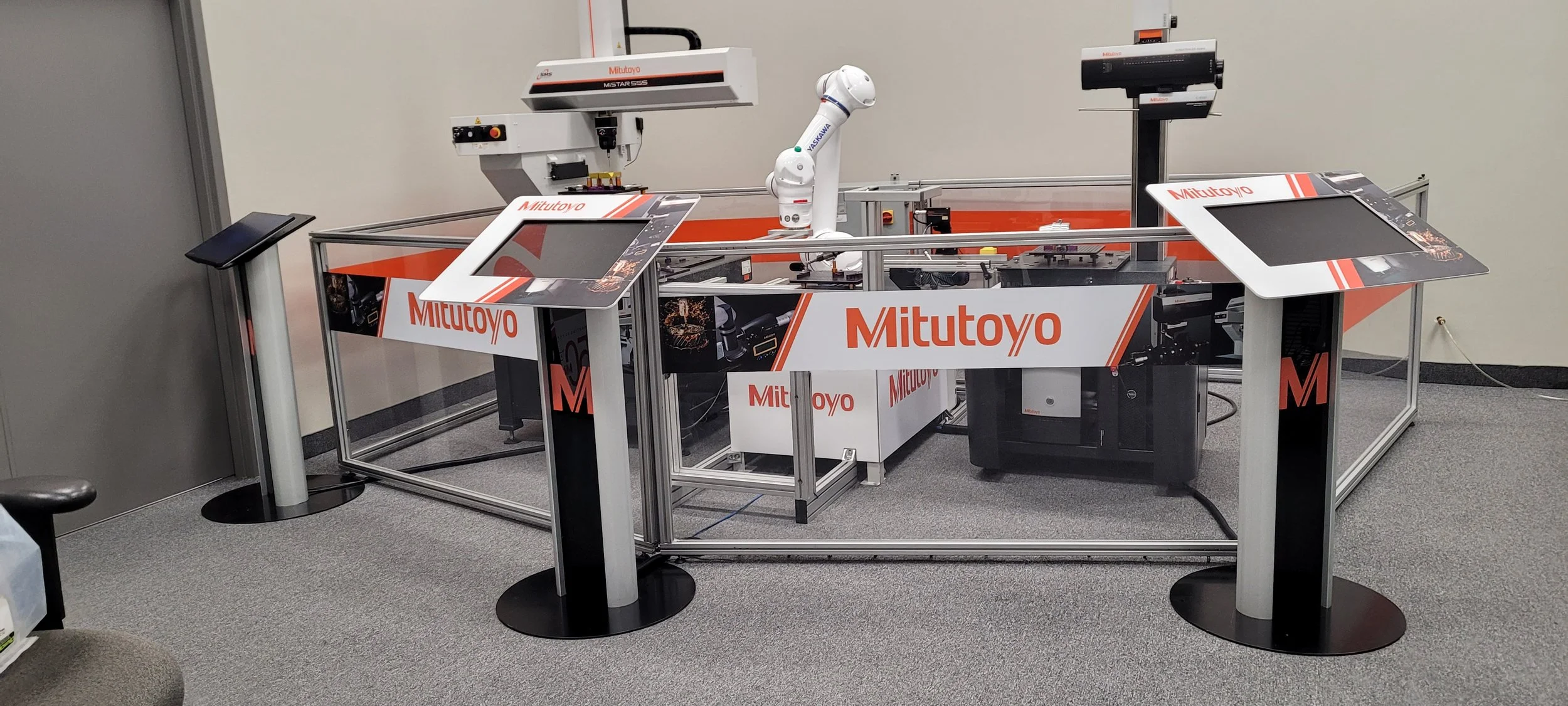 Industrial robotic arm and machinery inside a fenced area by Mitutoyo, with control screens on stands, in a lab or test environment.