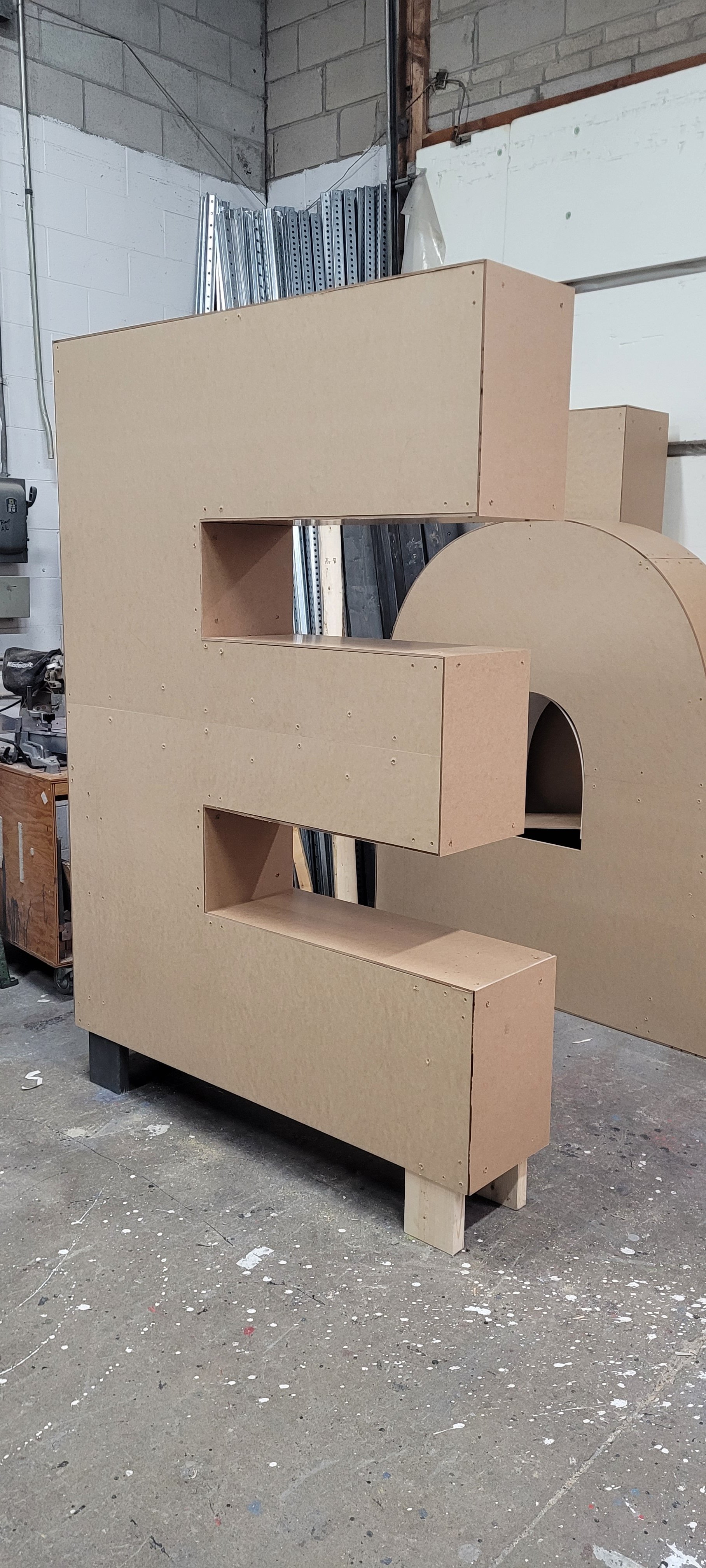Unfinished wooden furniture piece in a workshop, composed of multiple rectangular sections and a circular cutout.