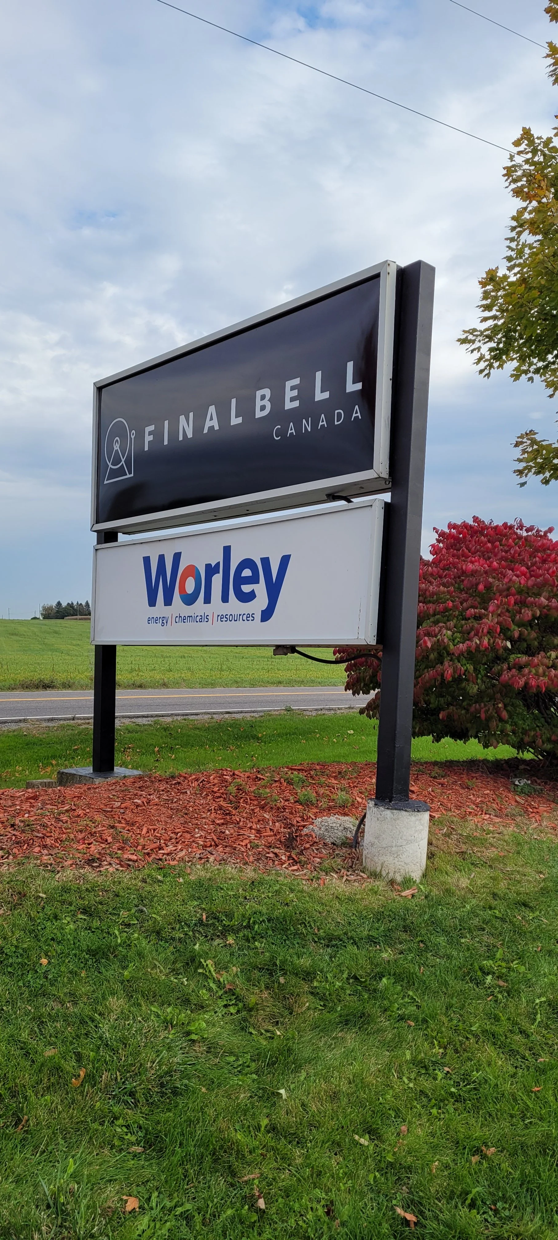 Outdoor signboard with two signs, one for Final Bell Canada and one for Worley, on a grassy area next to a road and a tree with red leaves.