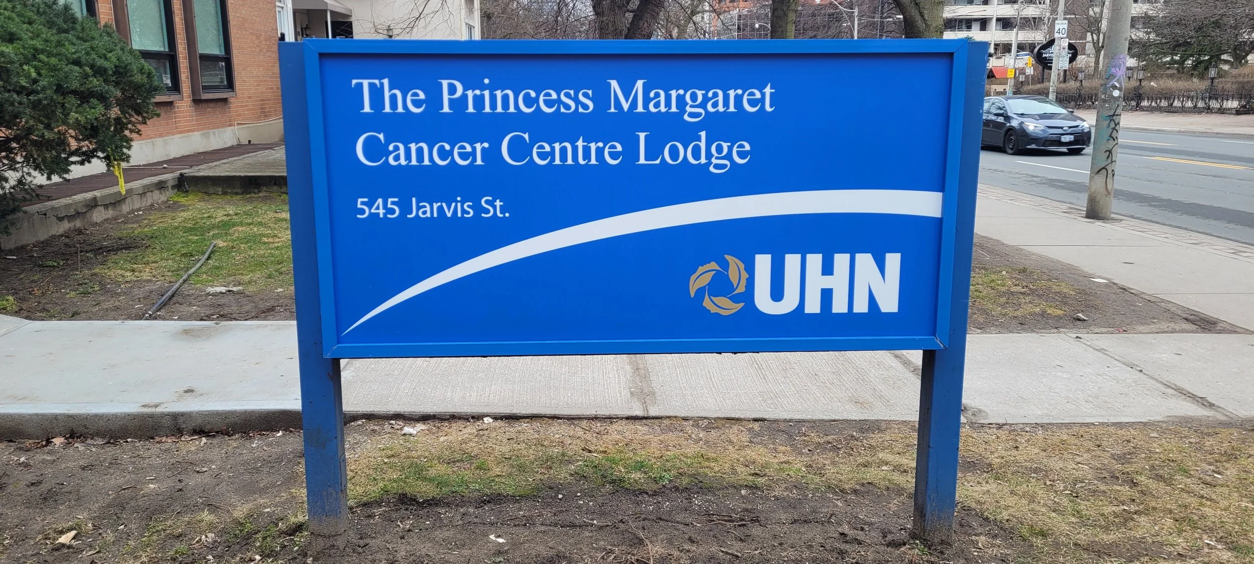 Blue sign for The Princess Margaret Cancer Centre Lodge at 545 Jarvis Street, with the UHN logo, on a sidewalk in an urban area.