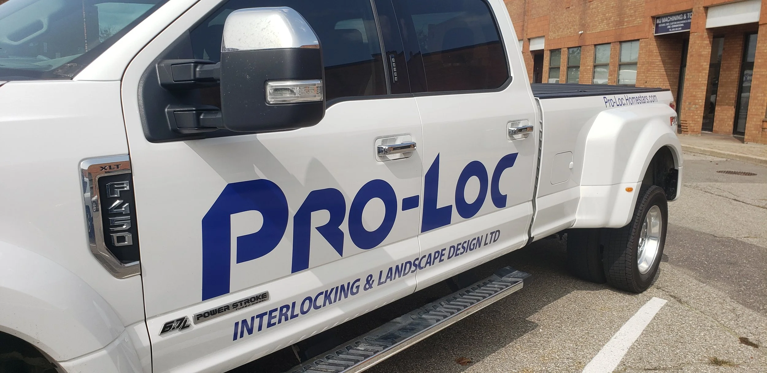 White pickup truck with Pro-Loc logos and text, parked on city street, with a brick building in the background.