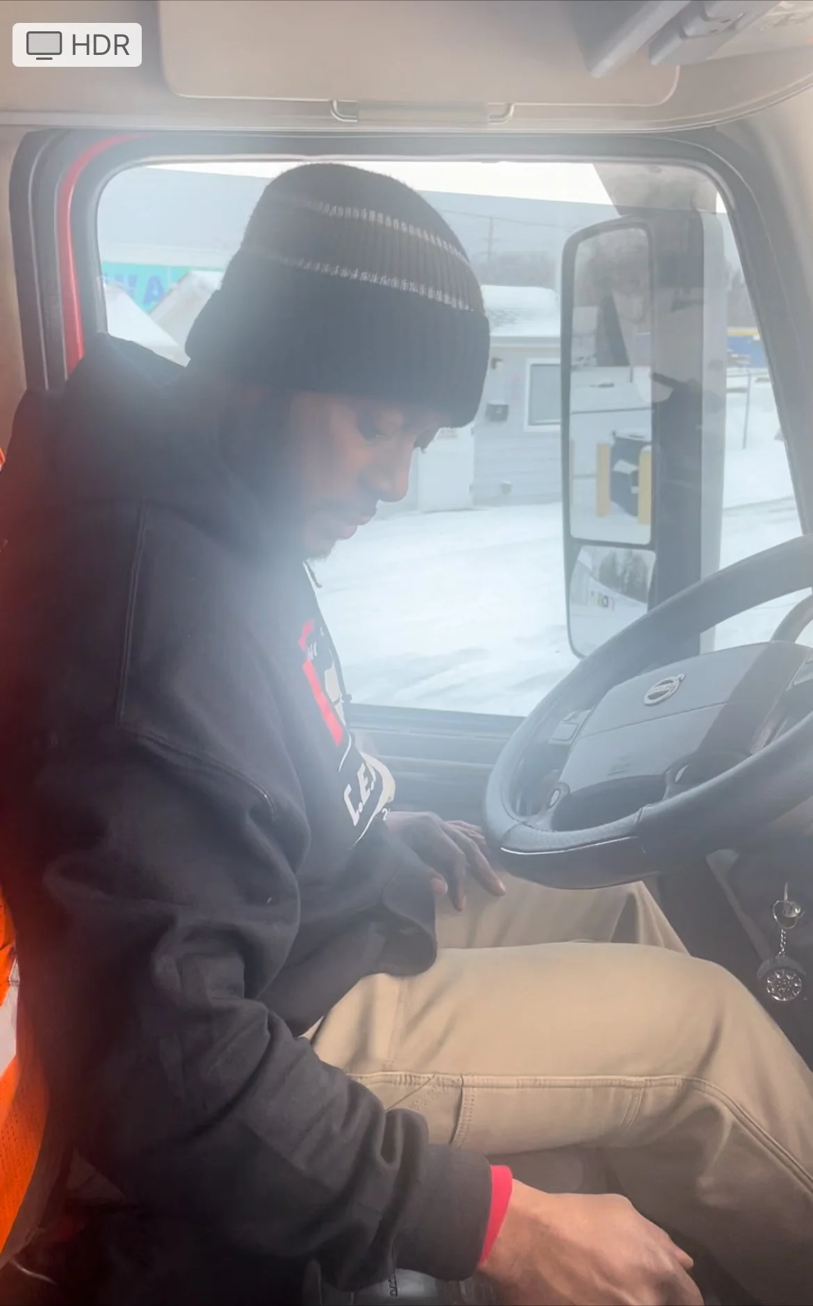 A man sitting inside a truck cabin, wearing a black beanie and black jacket, with snow visible outside through the windshield.